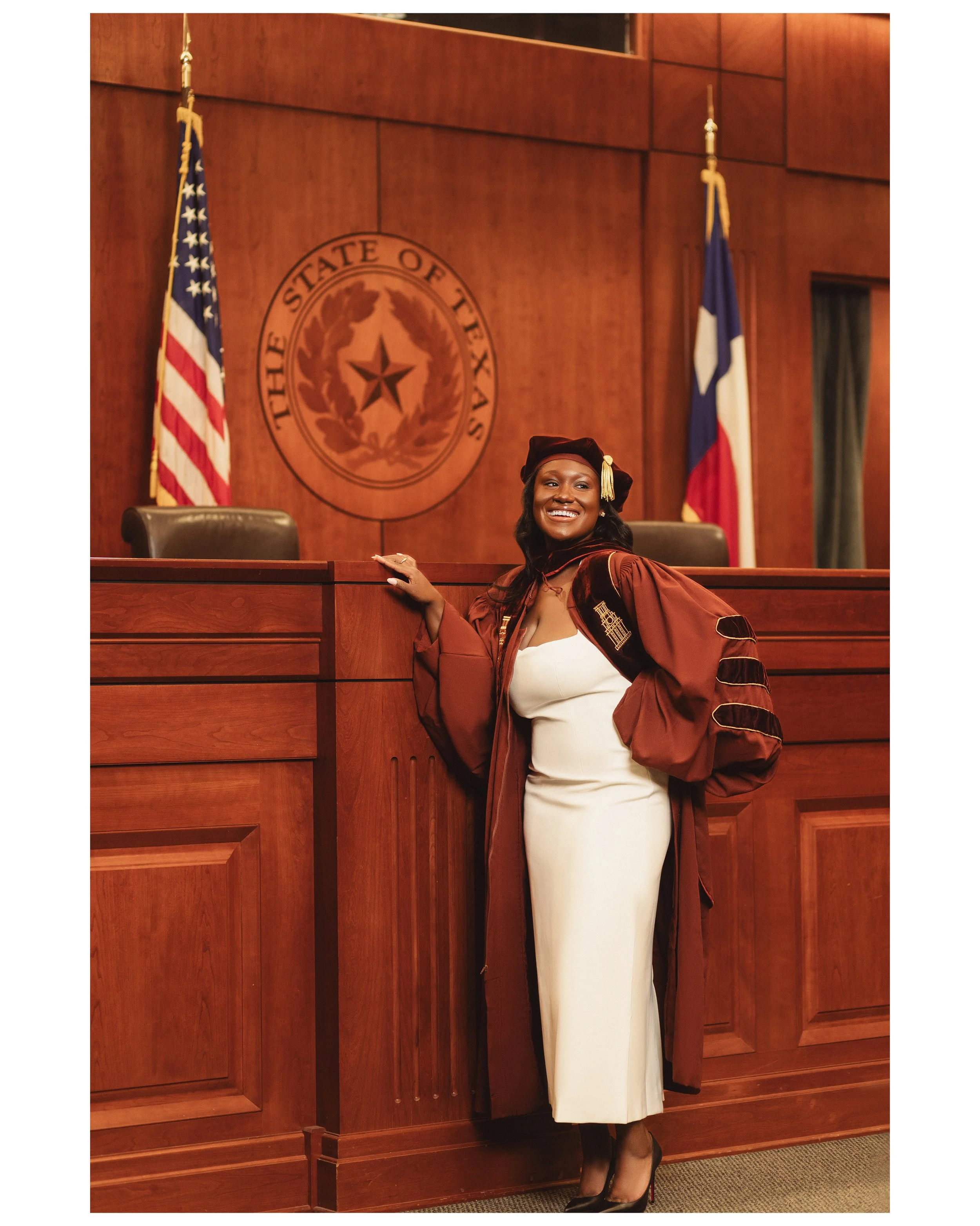 A woman in a graduation gown and cap standing in a courtroom with a wooden podium, American and Texas flags, and the Texas state seal in the background. photographed by Soba Grad, a graduation photography studio in Dallas, Texas