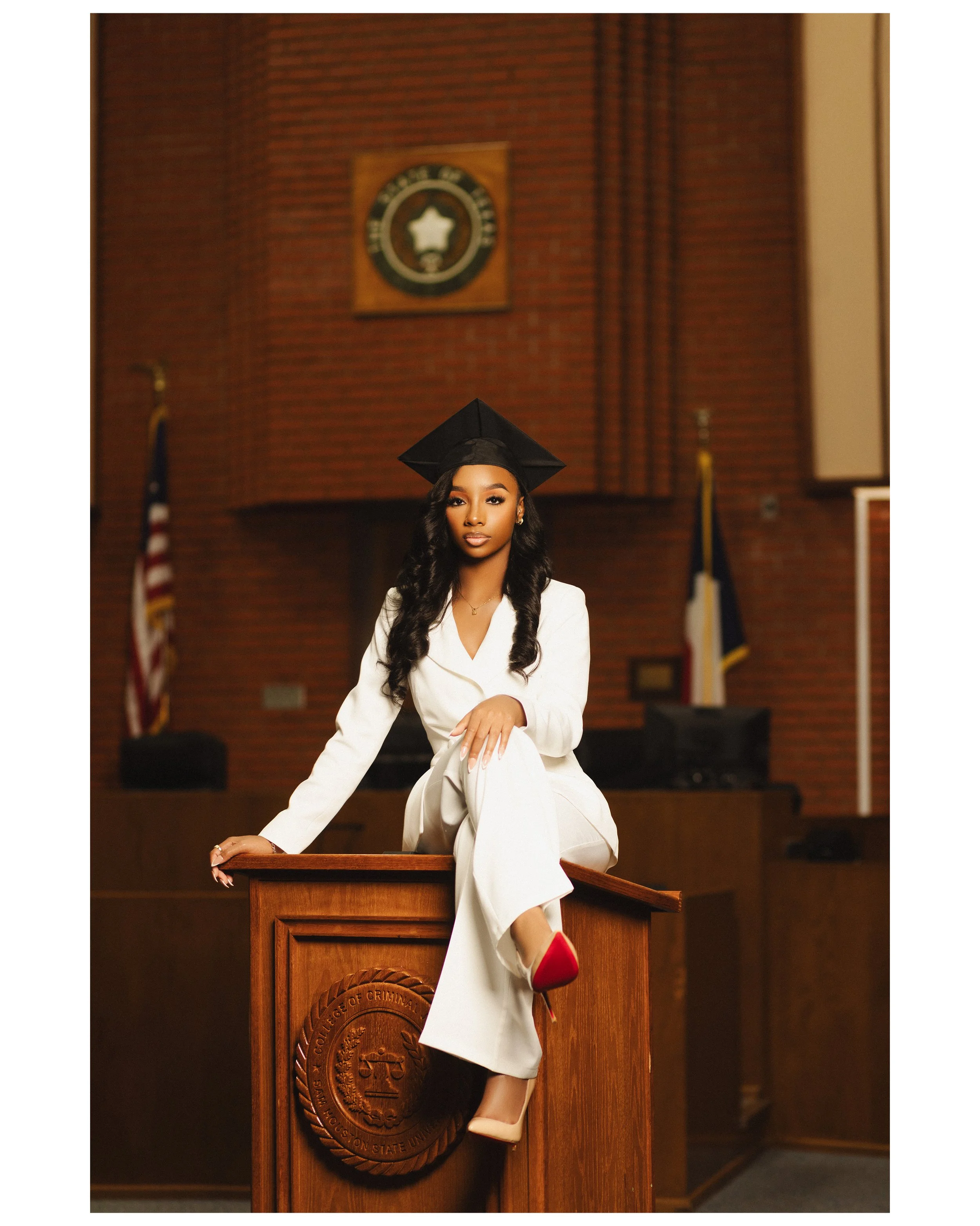 A young woman in a white suit and red heels, wearing a graduation cap, sitting on a judge's bench in a courtroom. photographed by Soba Grad, a graduation photography studio in Dallas, Texas