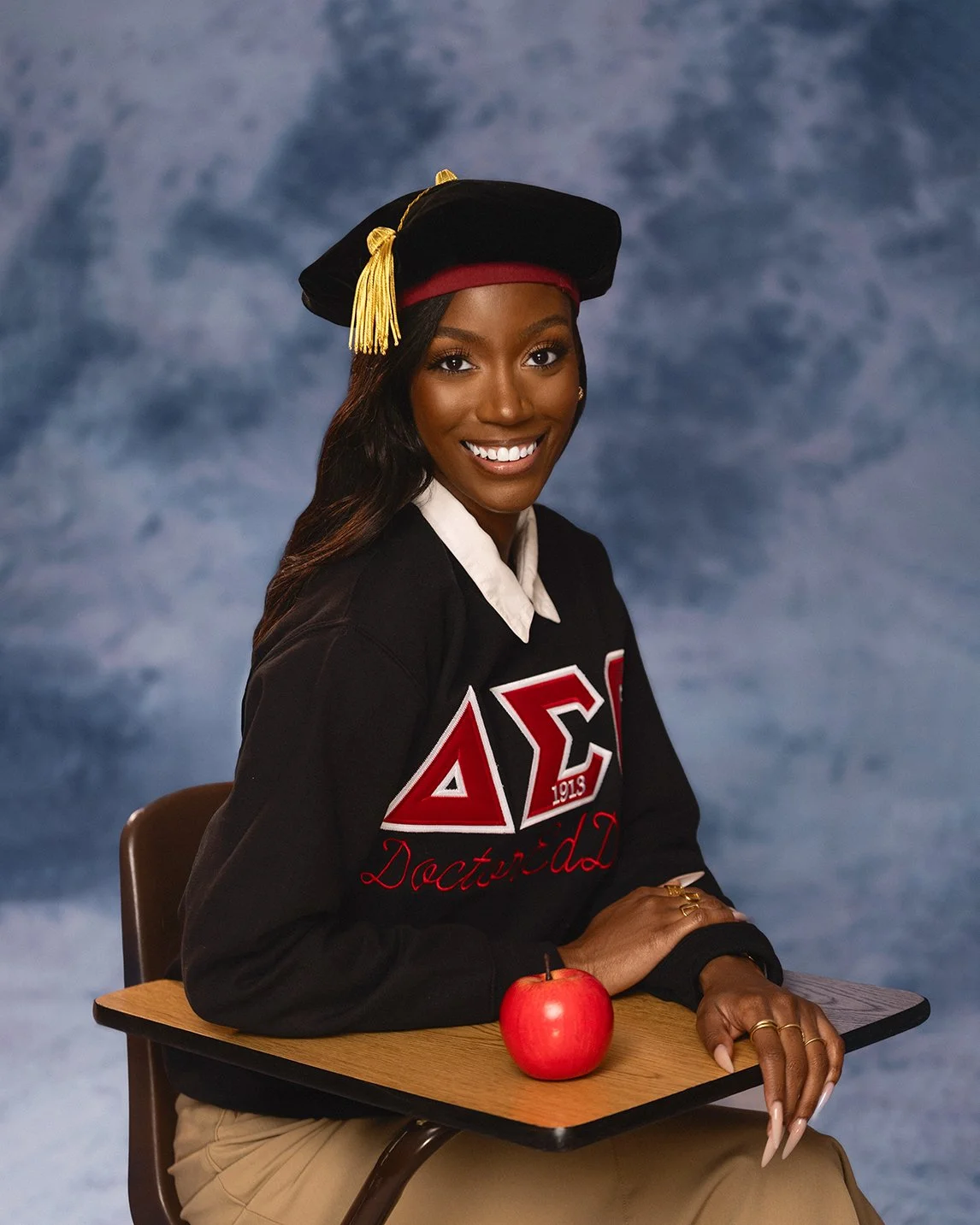A young woman wearing a black graduation cap and gown, sitting at a school desk with an apple, smiling at the camera, with a blue cloudy background.