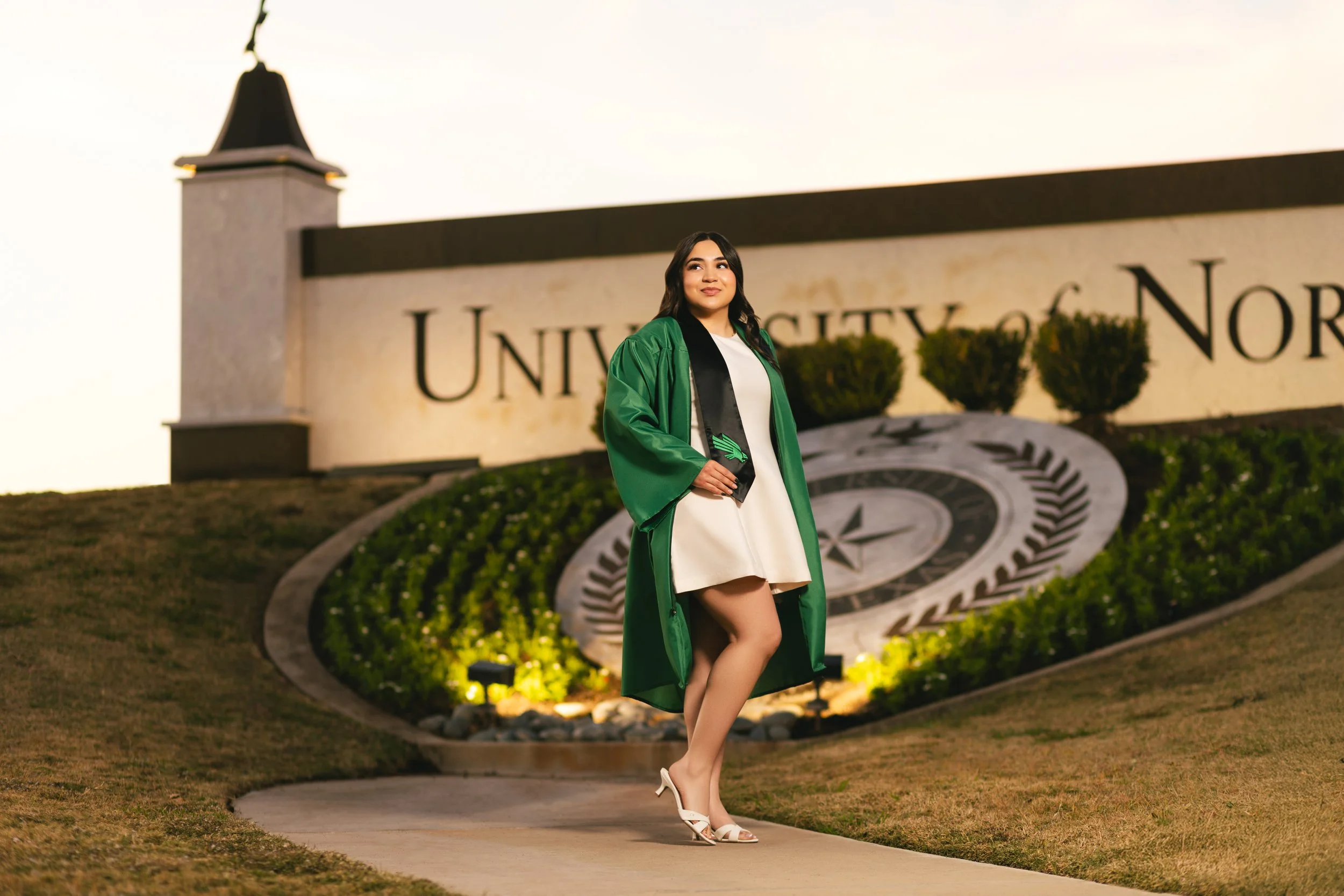 A woman in a green graduation gown and cap standing in front of a university sign during sunset. photographed by Soba Grad, a graduation photography studio in Dallas, Texas