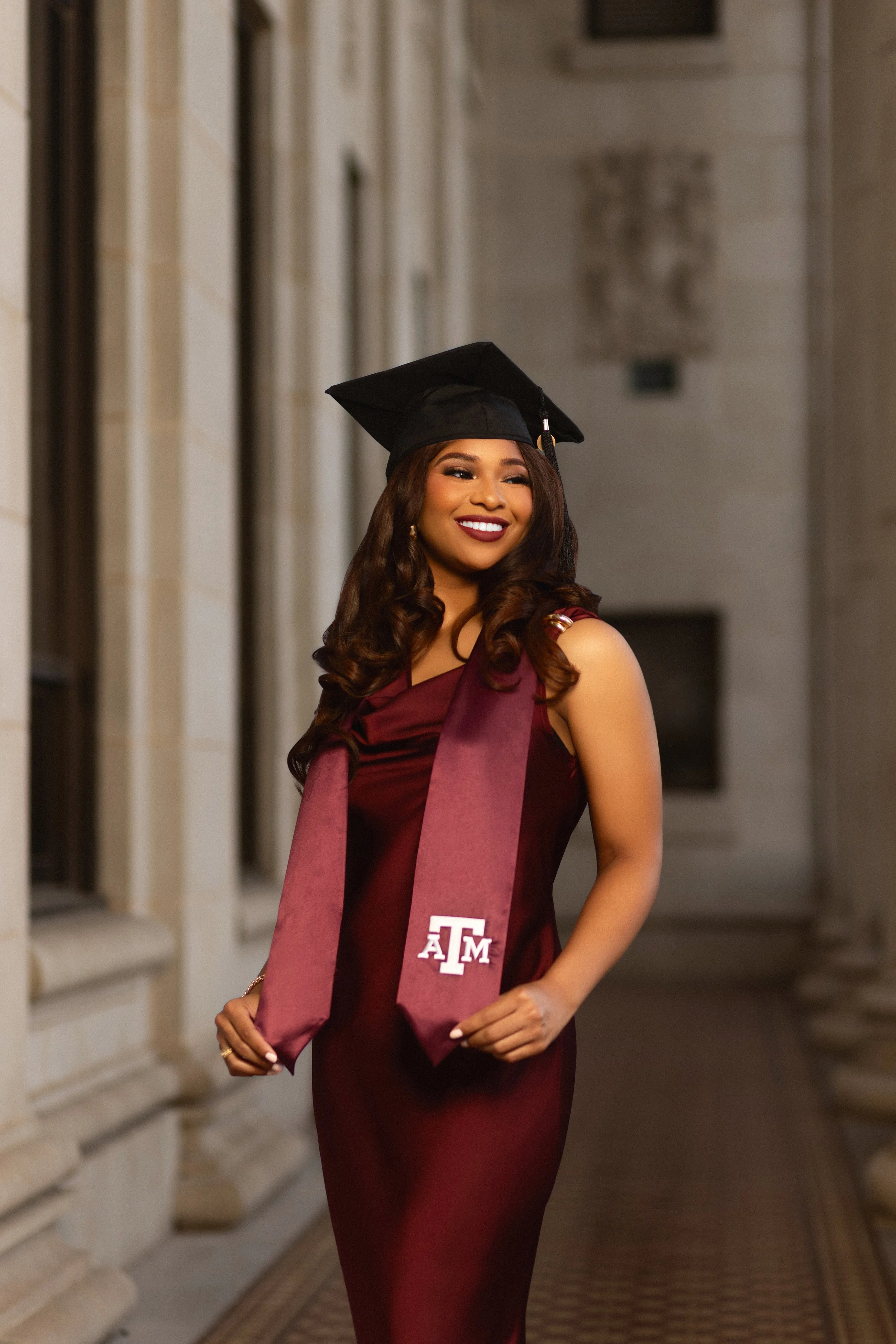 A young woman wearing a maroon graduation gown and cap, standing in a corridor with stone walls, smiling confidently. photographed by Soba Grad, a graduation photography studio in Dallas, Texas