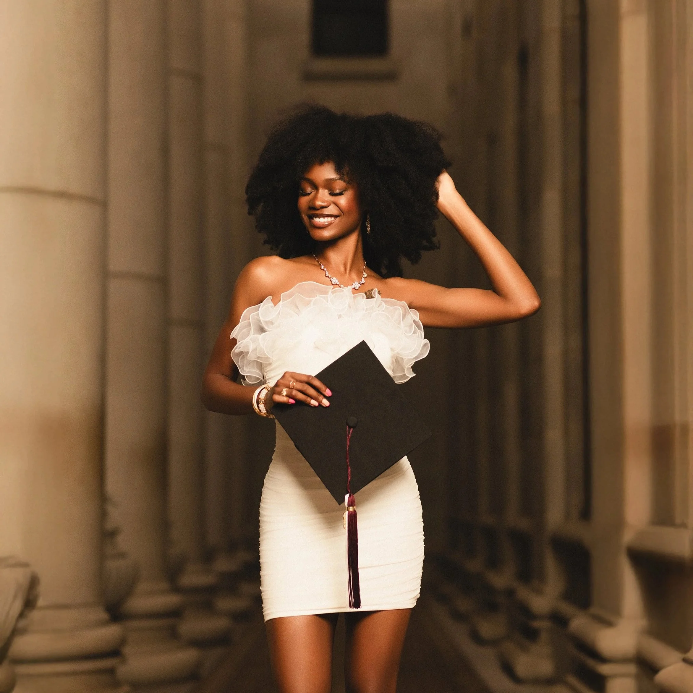 A smiling woman in a white dress holding a graduation cap in a corridor with columns. photographed by Soba Grad, a graduation photography studio in Dallas, Texas