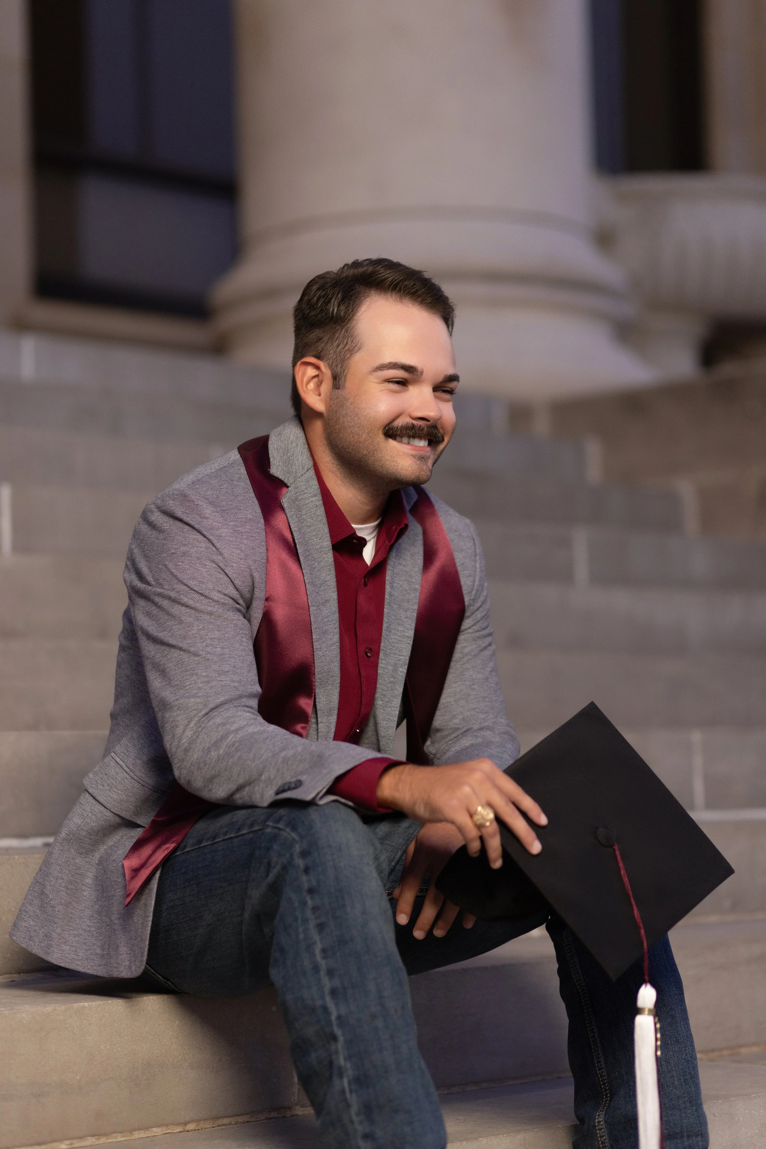A young man with a mustache and short brown hair, smiling, sitting on stairs outside a government or university building, holding a black graduation cap in his hand, dressed in a maroon shirt and gray jacket. photographed by Soba Grad, a graduation.