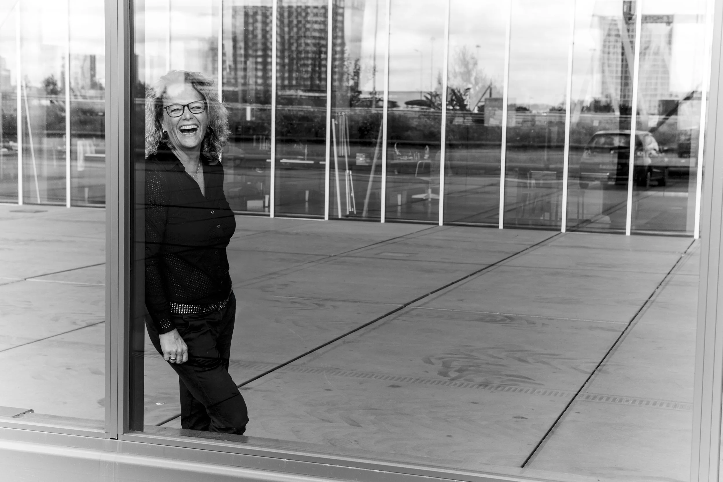 Black and white photo of a woman smiling near a glass wall, reflecting urban architecture.
