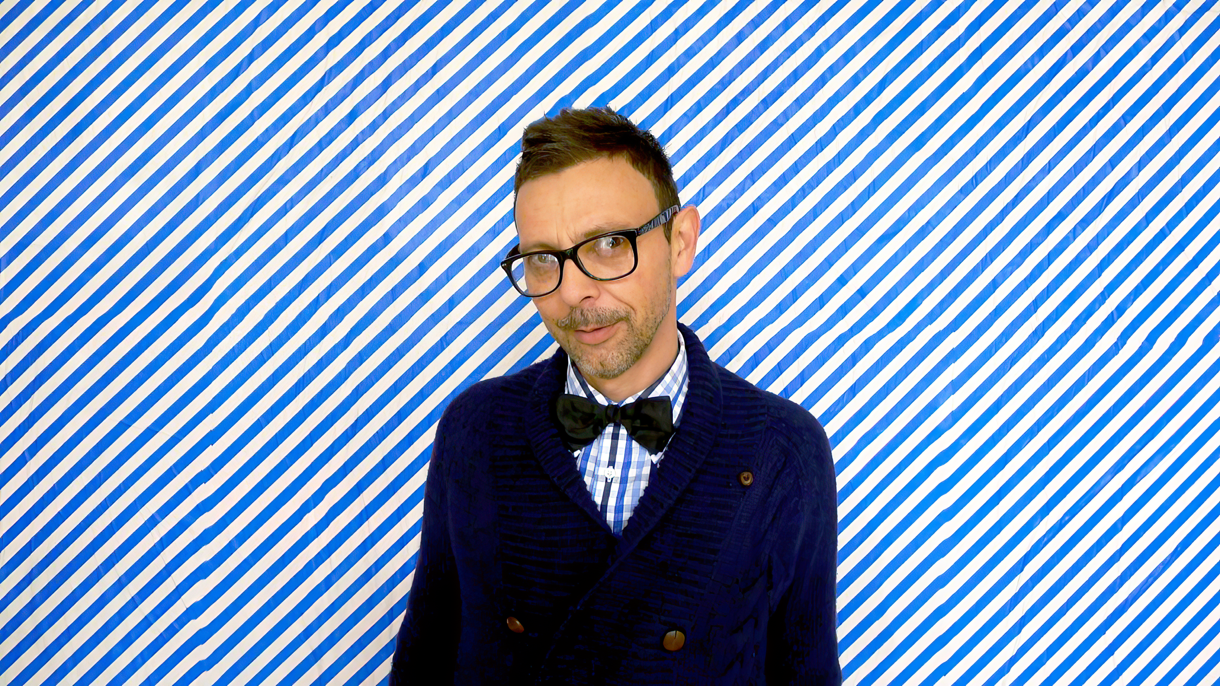 Un homme portant des lunettes, une chemise à carreaux, un nœud papillon noir et un cardigan bleu foncé pose devant un mur à rayures diagonales bleues et blanches.