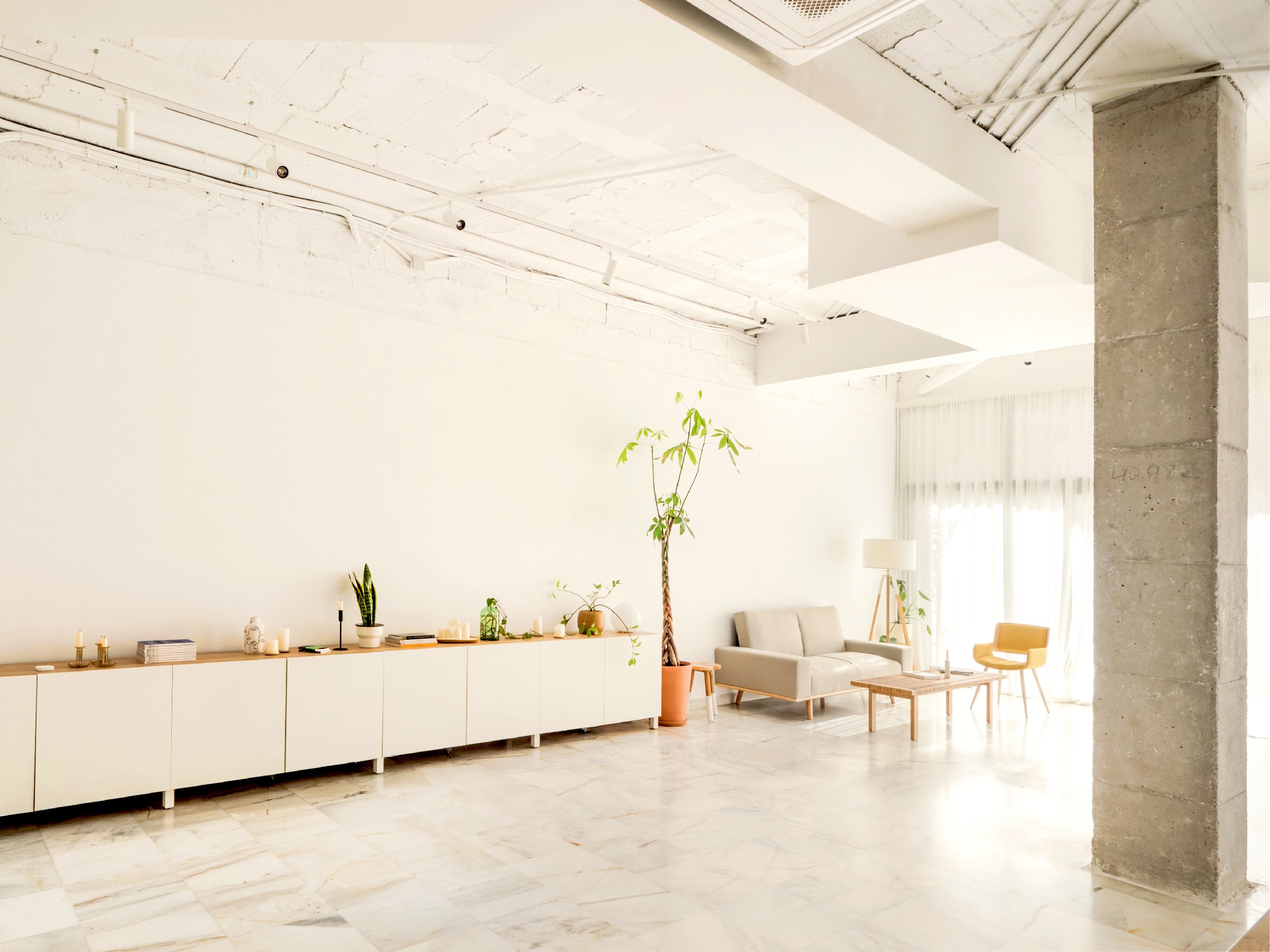 Espacio diáfano y despejado de 90 metros cuadrados con muebles modernos de color blanco, columnas de hormigón visto y techos altos de estilo industrial mediterráneo.