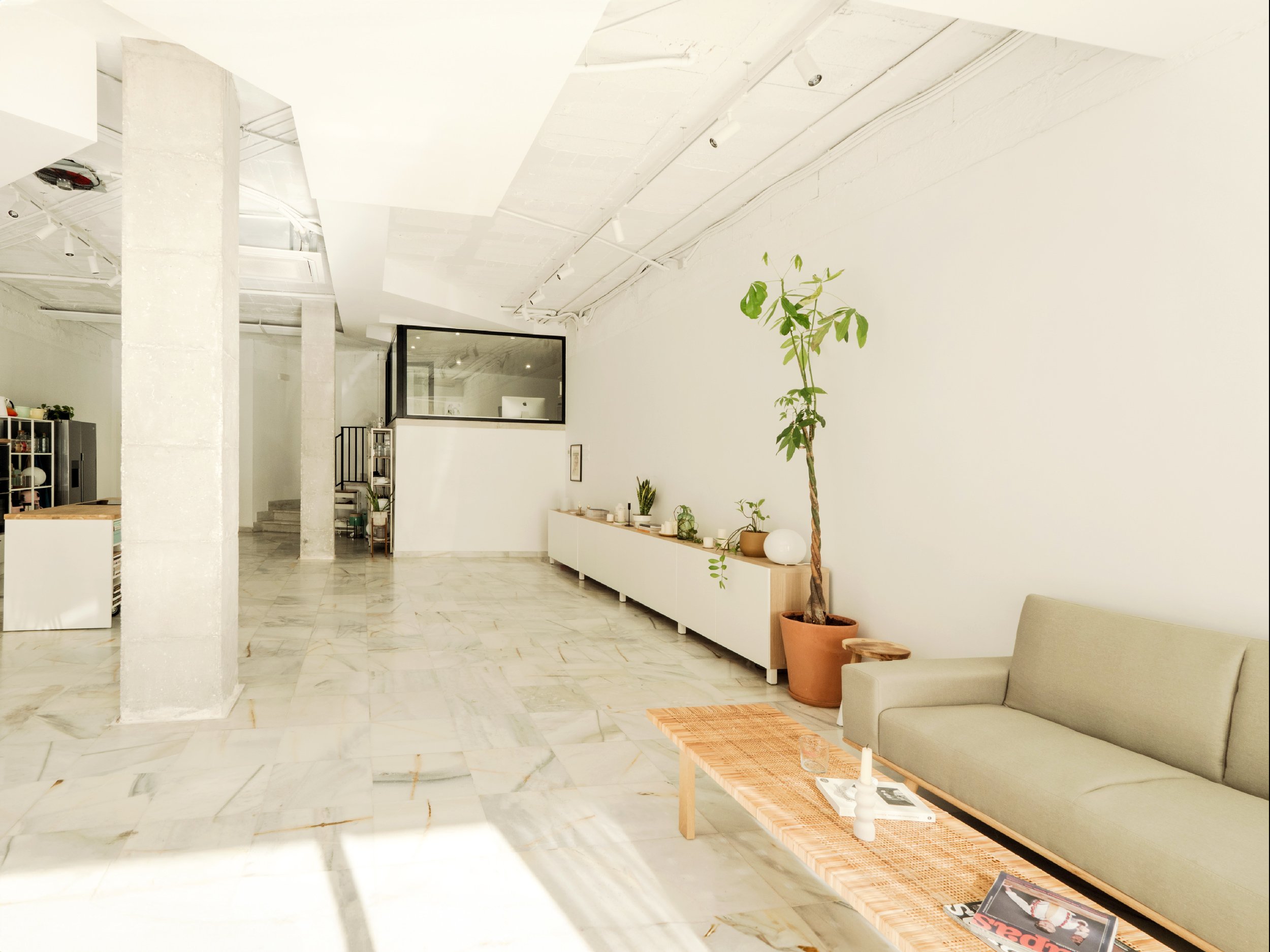 Sala amplia de 90 metros cuadrados con muebles modernos de color blanco, columnas de hormigón visto y techos altos de estilo industrial mediterráneo. Espacio privado de 22 m² insonorizado con una mampara corredera.