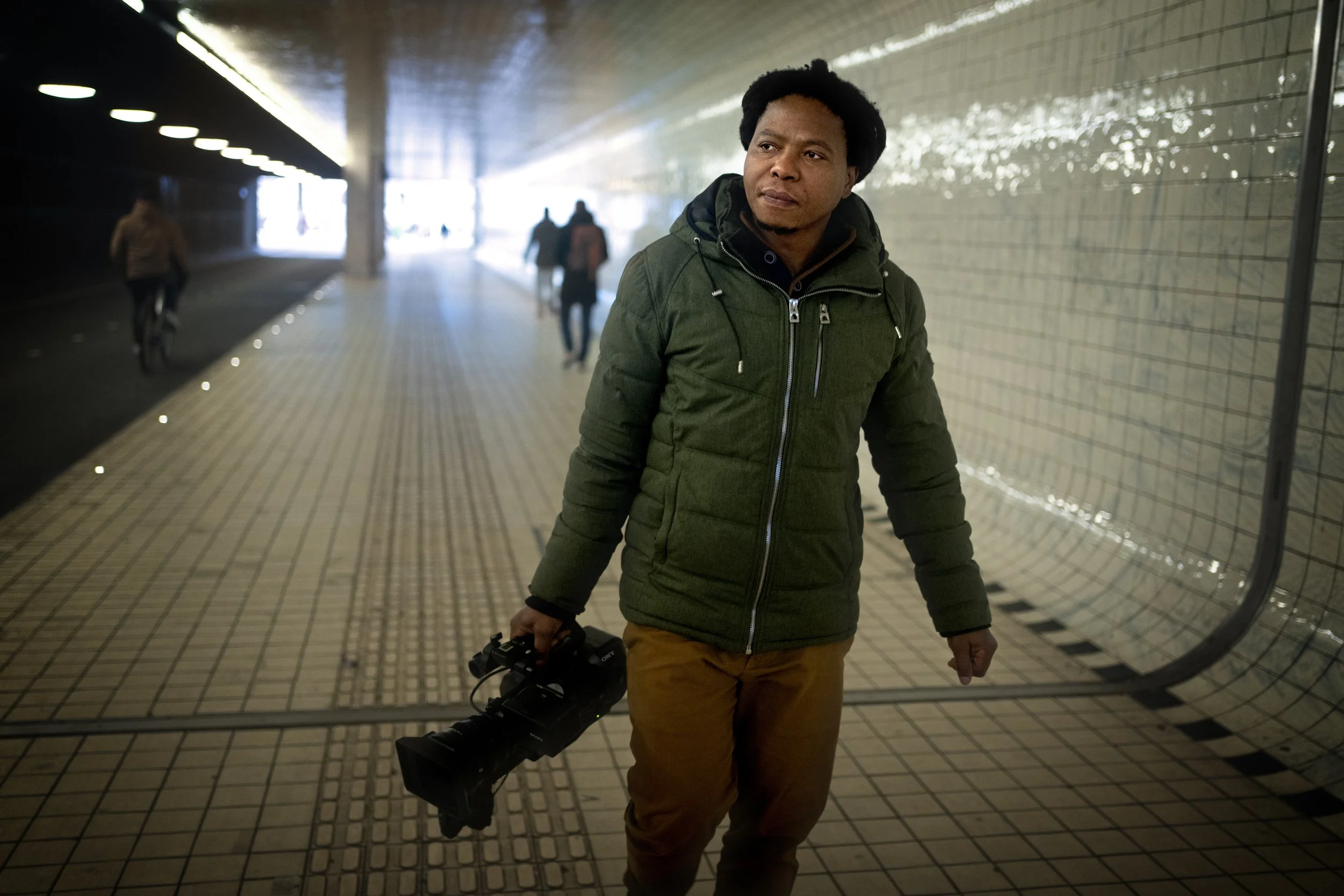 A man walking in an underground tunnel, carrying a professional video camera in his right hand, with a few pedestrians in the background.