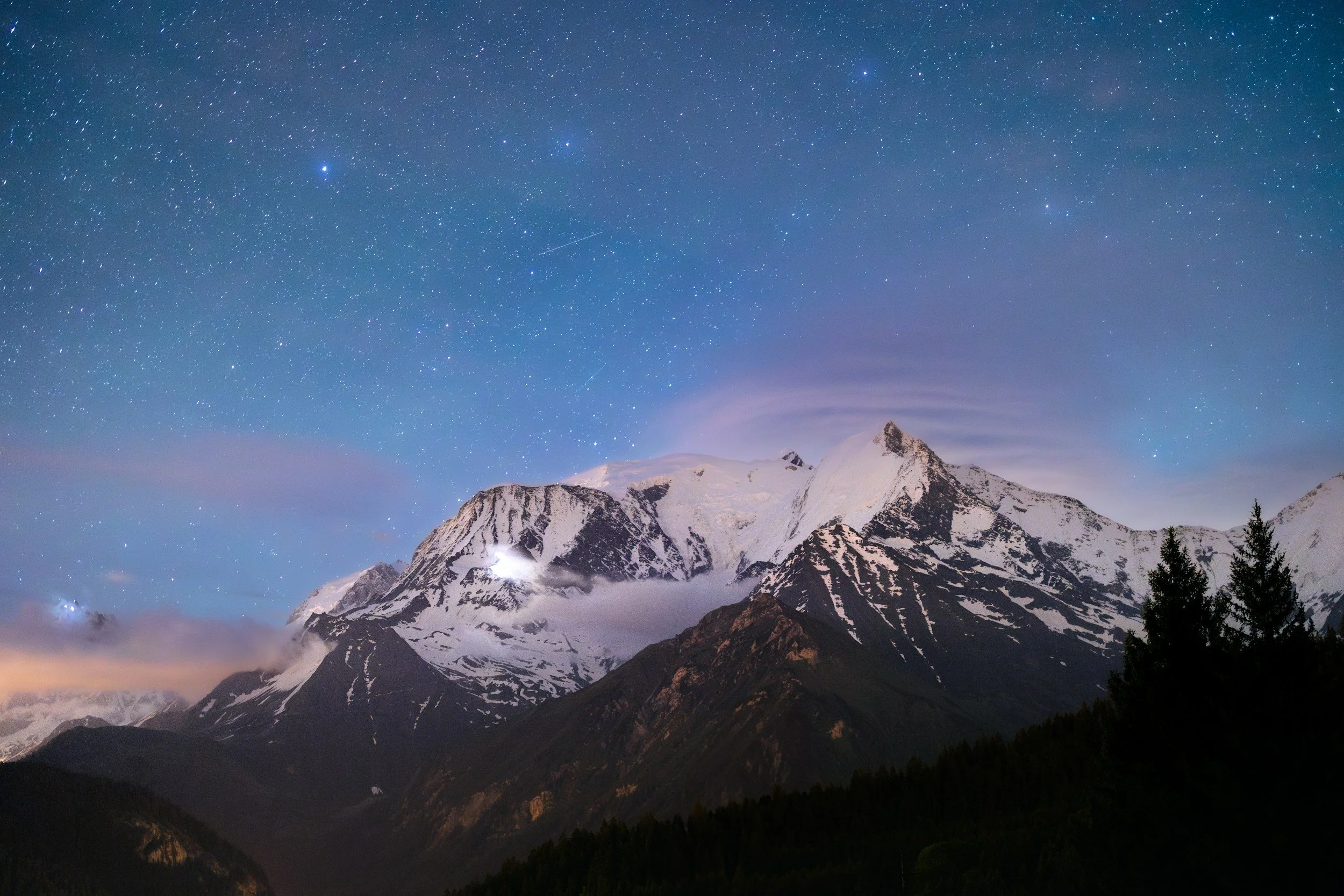 Snow-capped mountains under a starry night sky with clouds and trees in the foreground.