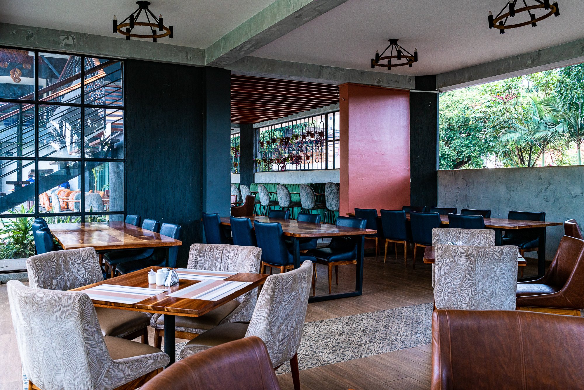 Interior of a modern restaurant with wooden tables, various patterned chairs, large window with greenery outside, hanging lights, and decorative wall art.