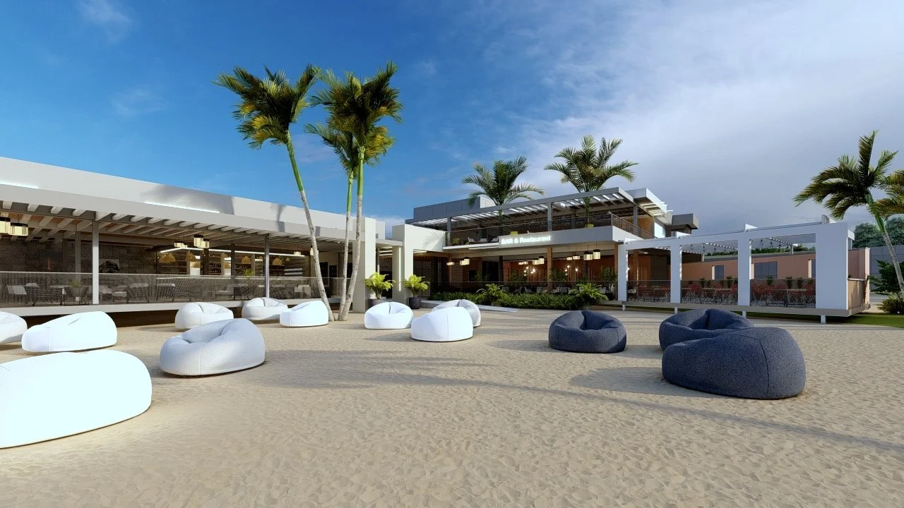 A beachside outdoor lounge area with white and dark blue bean bag chairs, palm trees, a sandy surface, and a modern building in the background with a bar and restaurant.