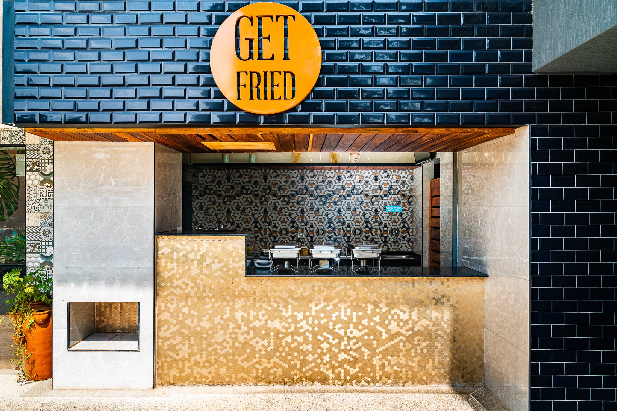 A food stand with a sign that says "Get Fried". The stand has a black tiled exterior, a gold mosaic front, and a patterned tile background behind the serving window. Inside the stand, there are three chafing dishes, and to the side, some plants are v