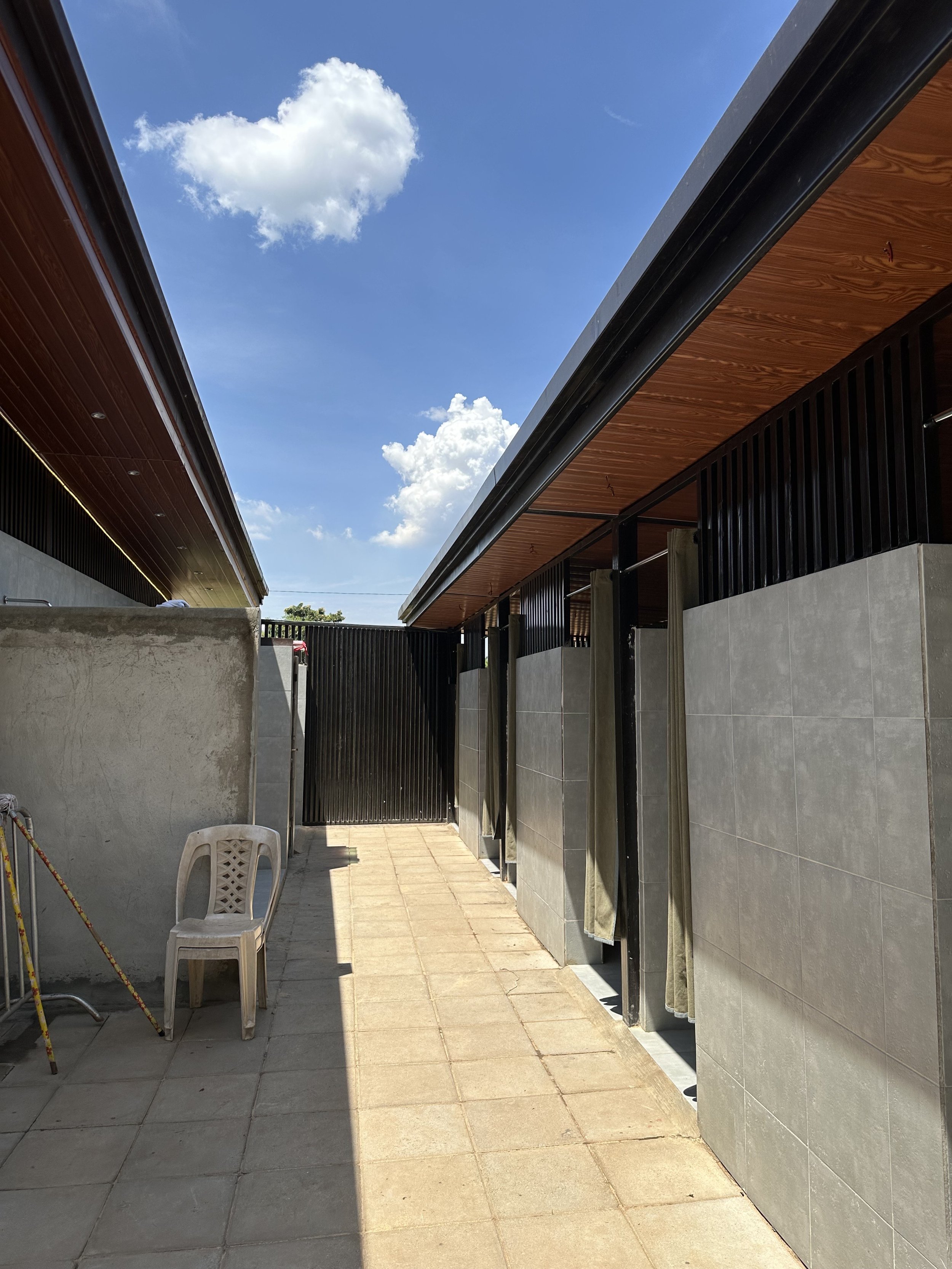 An outdoor shower area with a tiled floor, grey walls, and showers with curtains, under a partly cloudy blue sky.