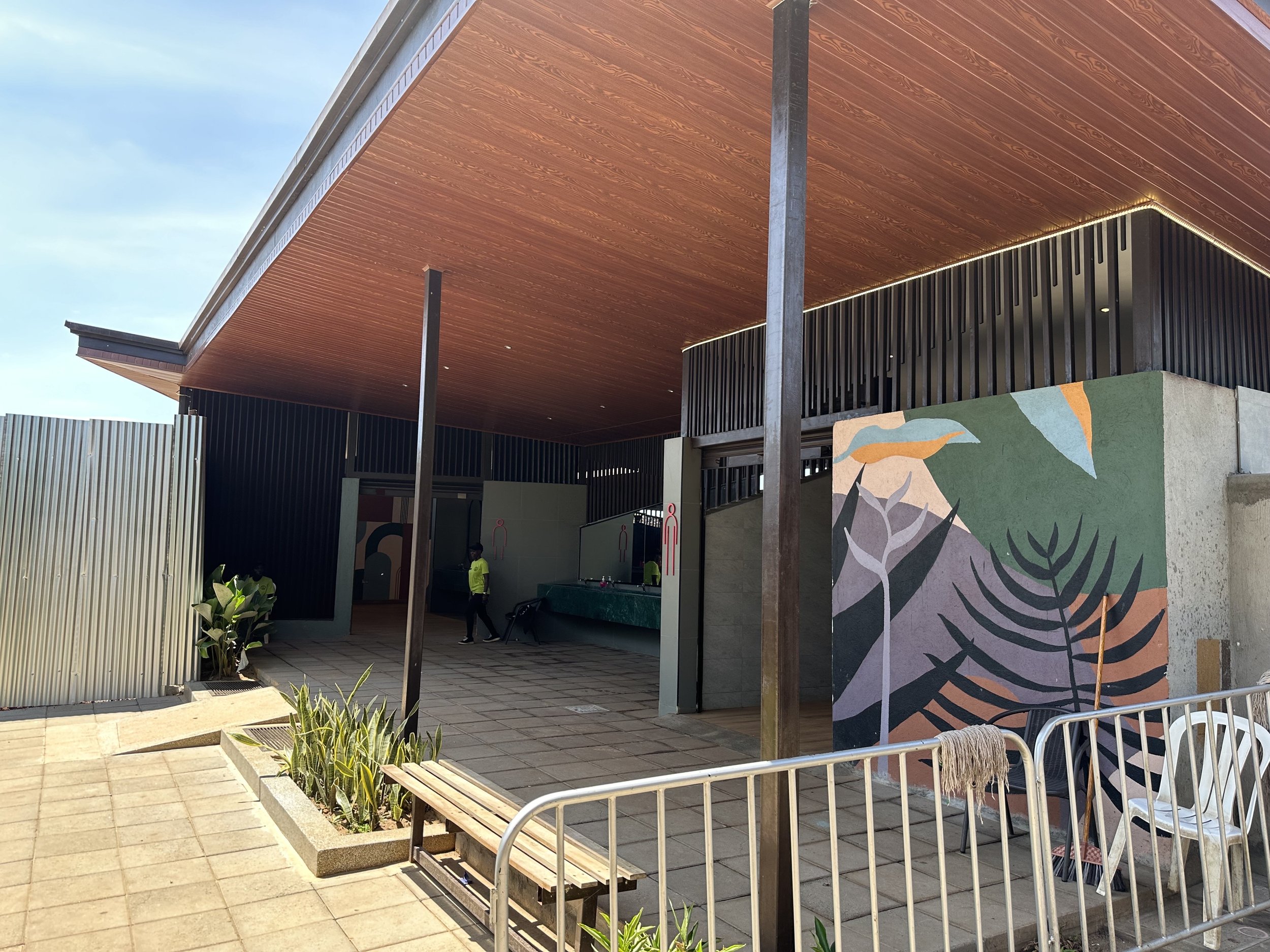 Exterior of a modern building with a wooden roof, black vertical slats, a colorful mural with abstract plant designs, and a small outdoor area with plants, benches, and barriers.