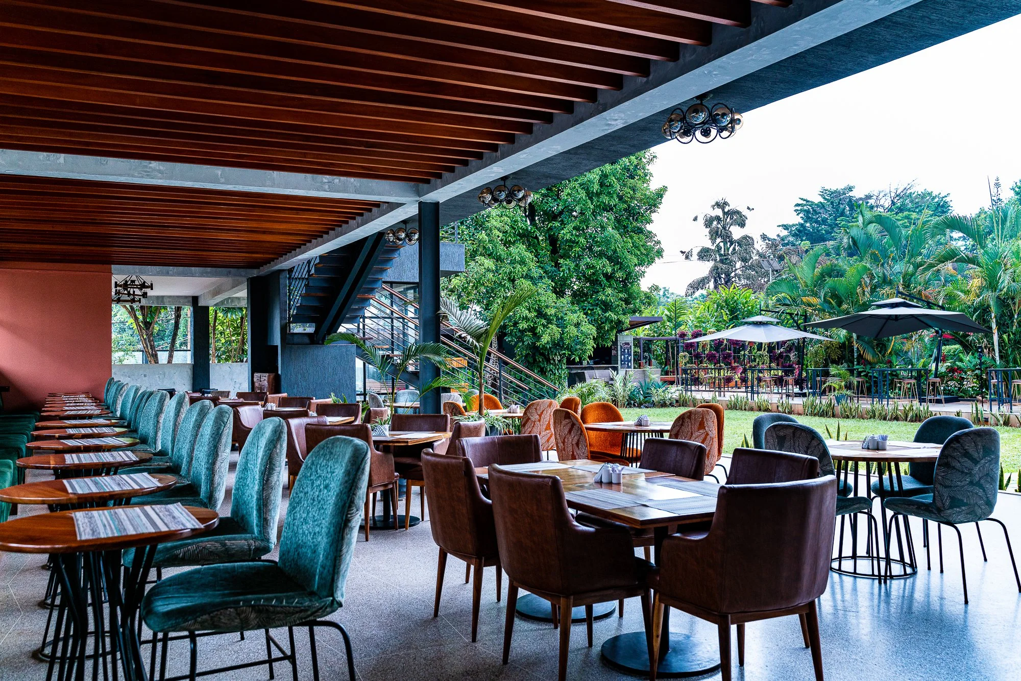 Indoor-outdoor restaurant area with wooden ceiling, colorful chairs, and a view of lush greenery and outdoor umbrellas.