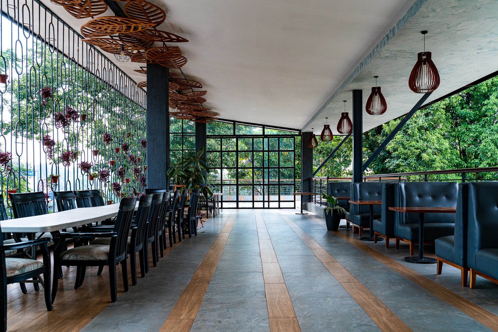 Interior of a modern restaurant or cafe with a long white table and black chairs on the left, and blue booth seating with small tables on the right, with large windows overlooking greenery, wooden and gray tiled flooring, and decorative hanging light
