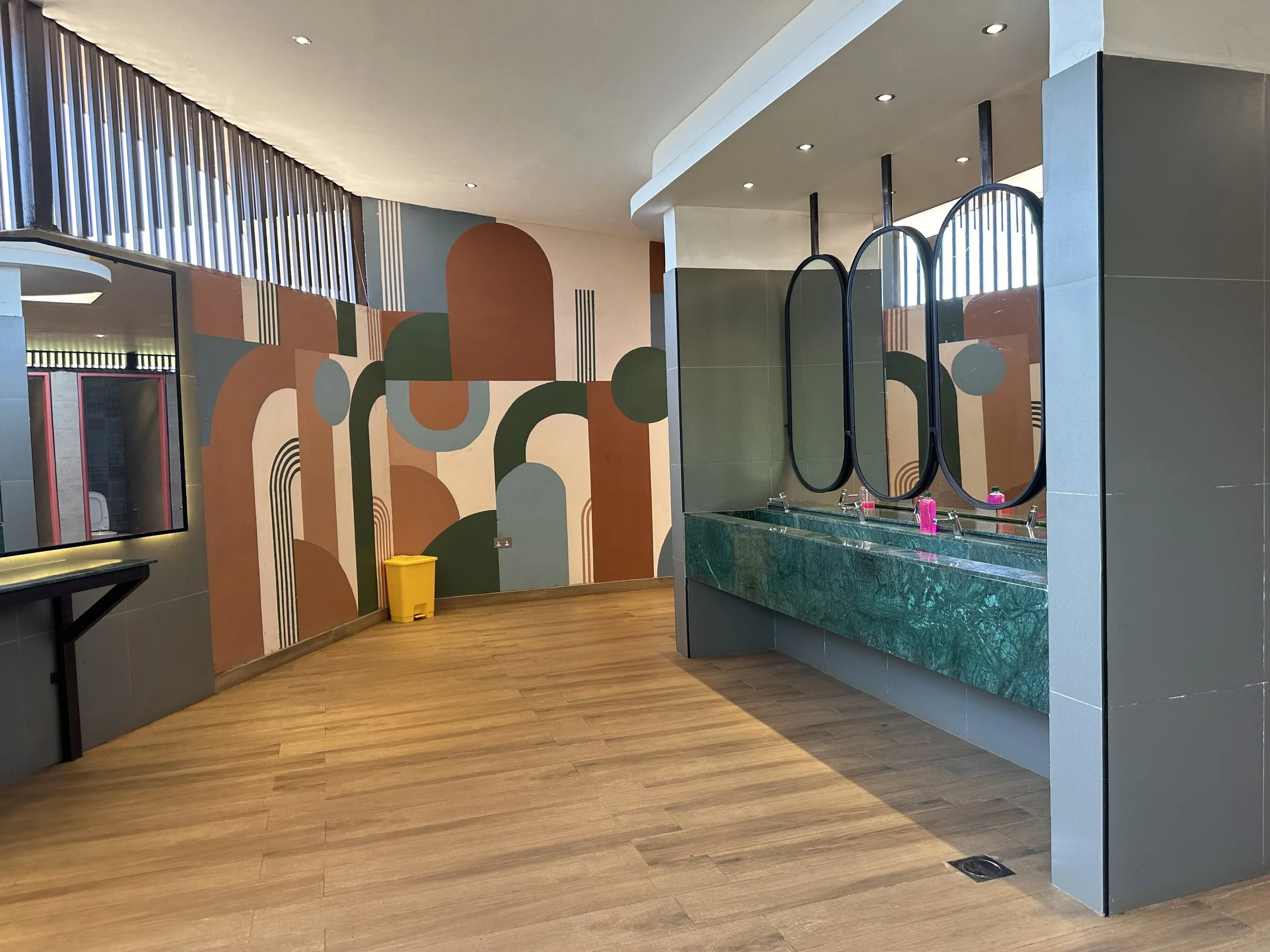 Public restroom with a green marble sink, three mirrors with black frames, and a colorful abstract mural on the wall.