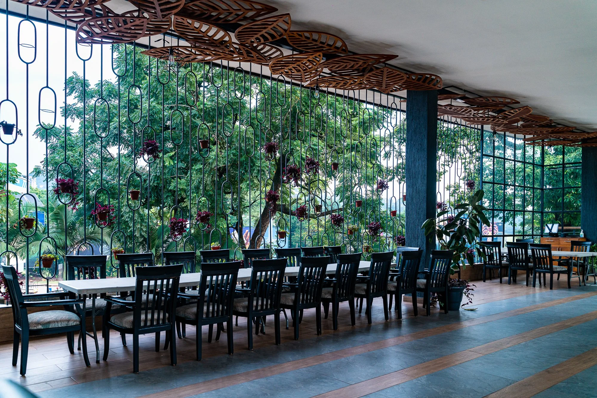 Interior of a restaurant with a long white table, black chairs, and decorative wooden leaf patterns hanging from the ceiling. Large windows with greenery outside and hanging flower pots.