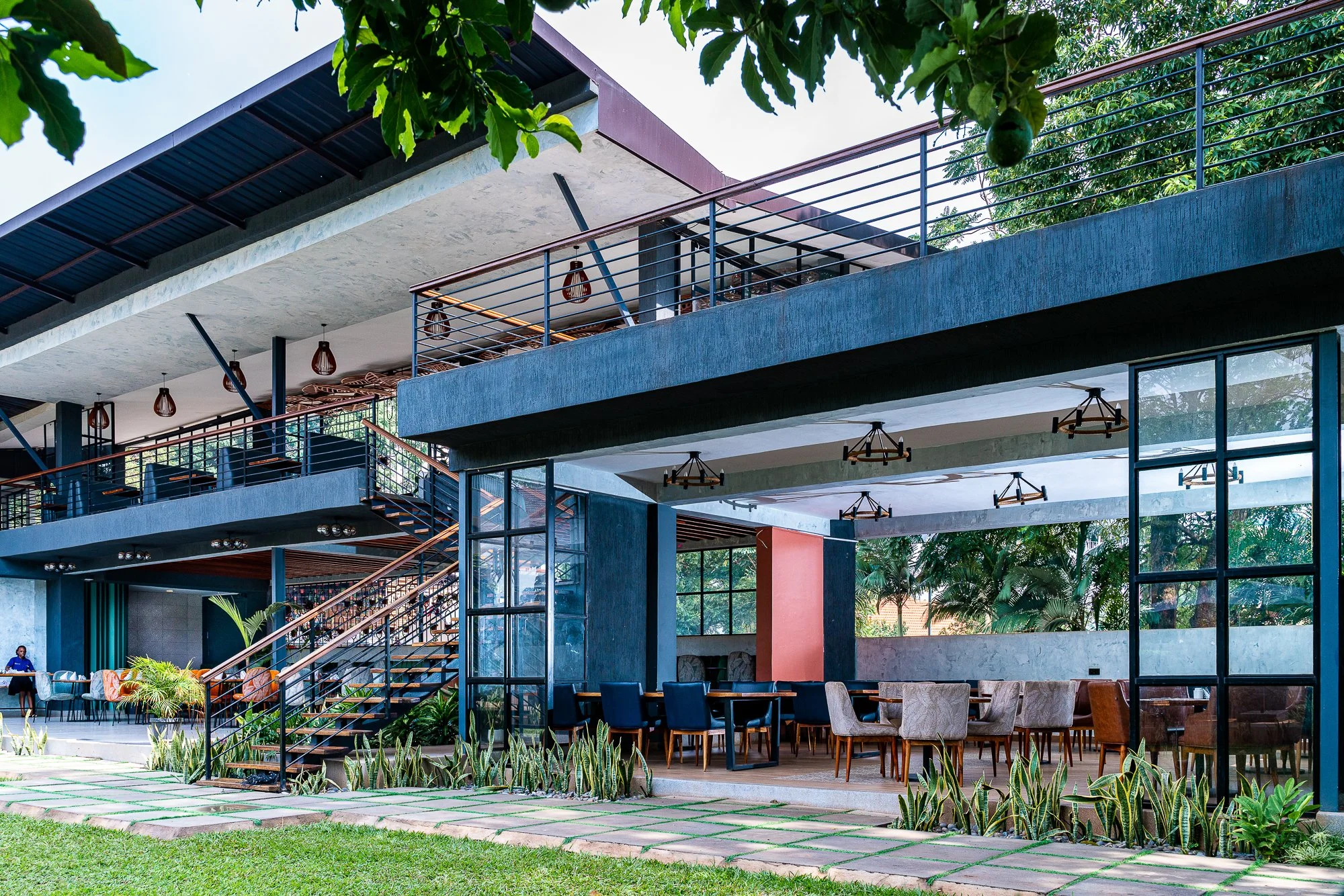 Modern multi-level building with outdoor dining area, glass walls, and greenery.