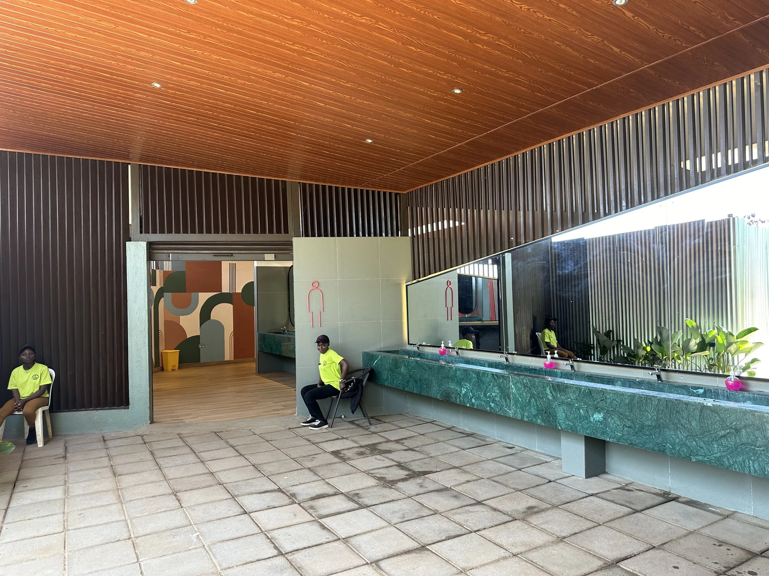 An indoor space with a modern design, featuring a green marble counter, large mirror, and wooden ceiling. There are three people sitting and waiting, two women wearing bright yellow shirts, one sitting on a chair near the wall and the other on a benc
