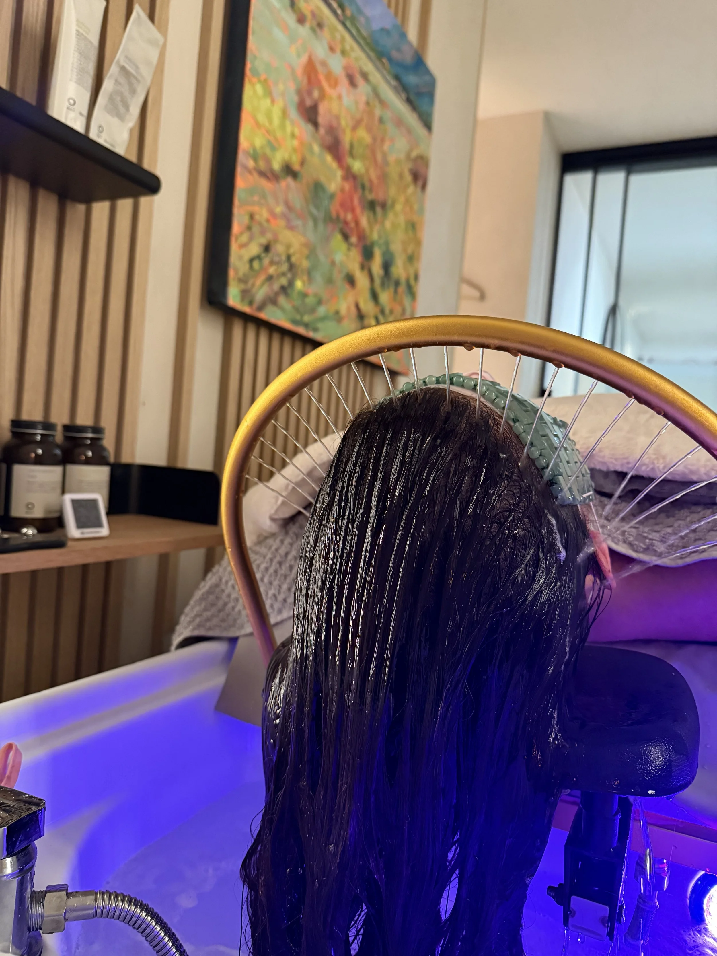 Person receiving a hair treatment in a salon, with hair sectioned and covered with a green cap at the scalp, sitting under a hair steamer with purple and blue lighting.