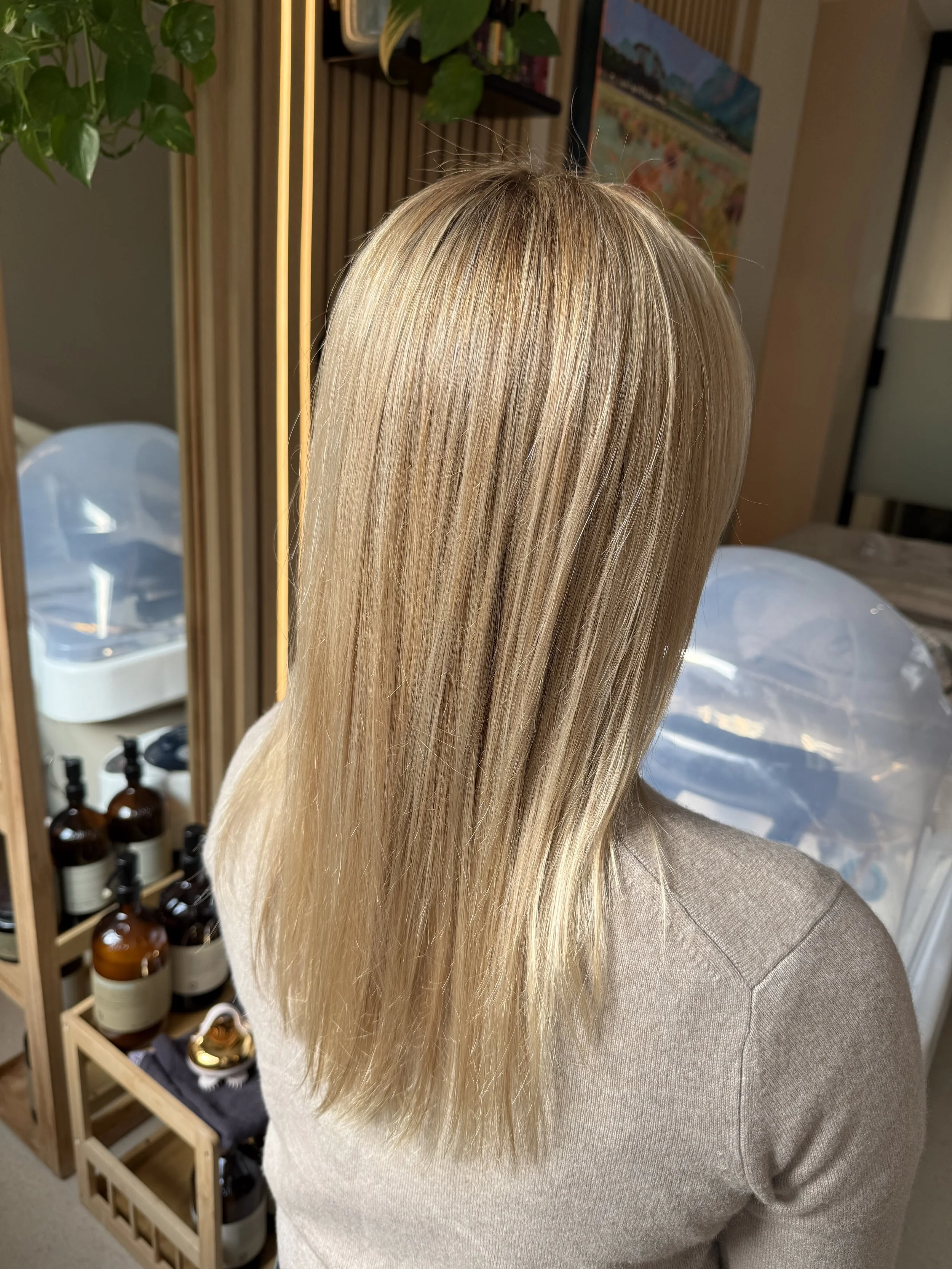 Blonde woman with shoulder-length wavy hair sitting in a salon chair, with a background of shelves, plants, and a couch.