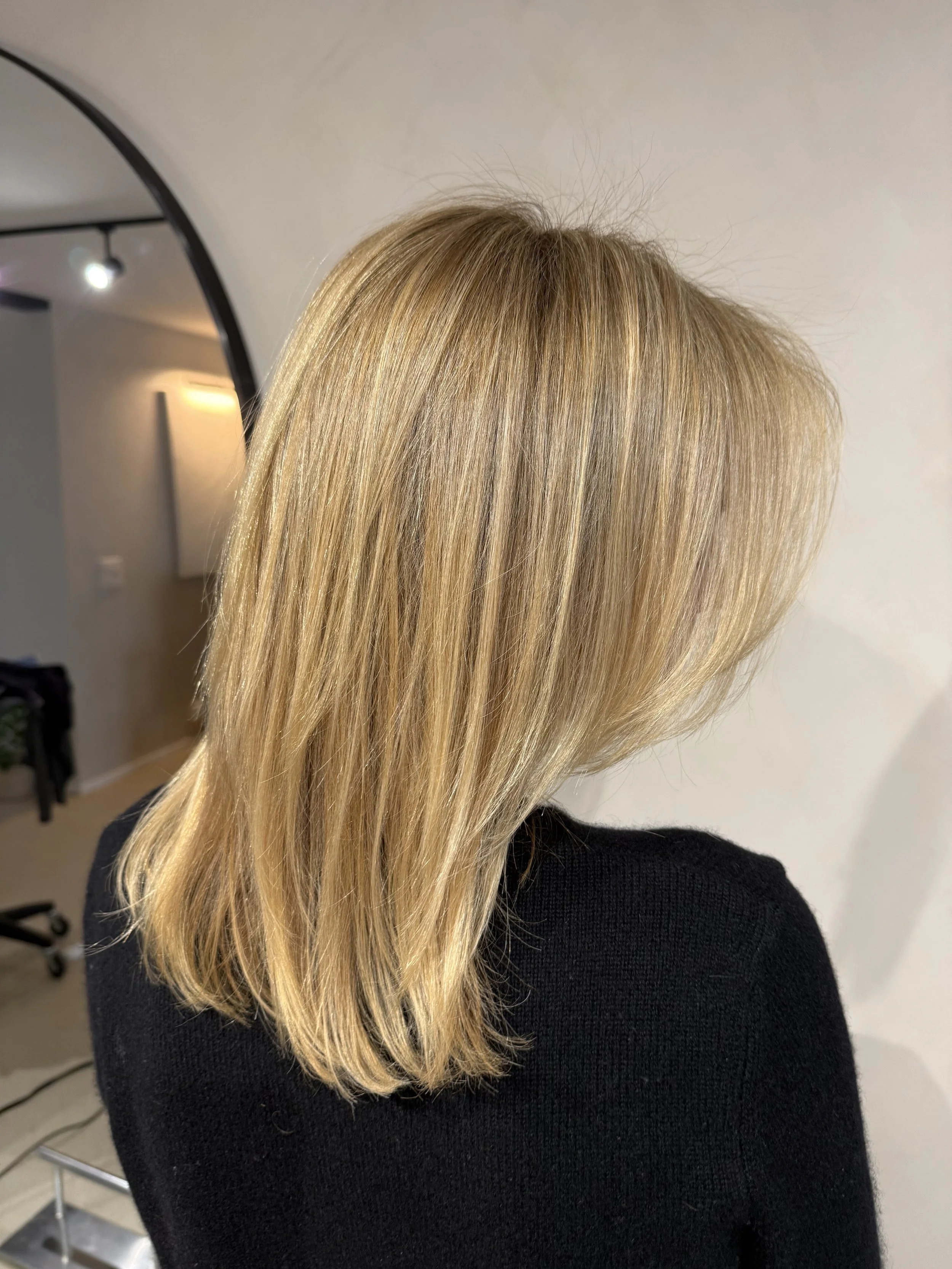 Side view of a woman with shoulder-length, wavy blonde hair with subtle highlights in a salon setting.