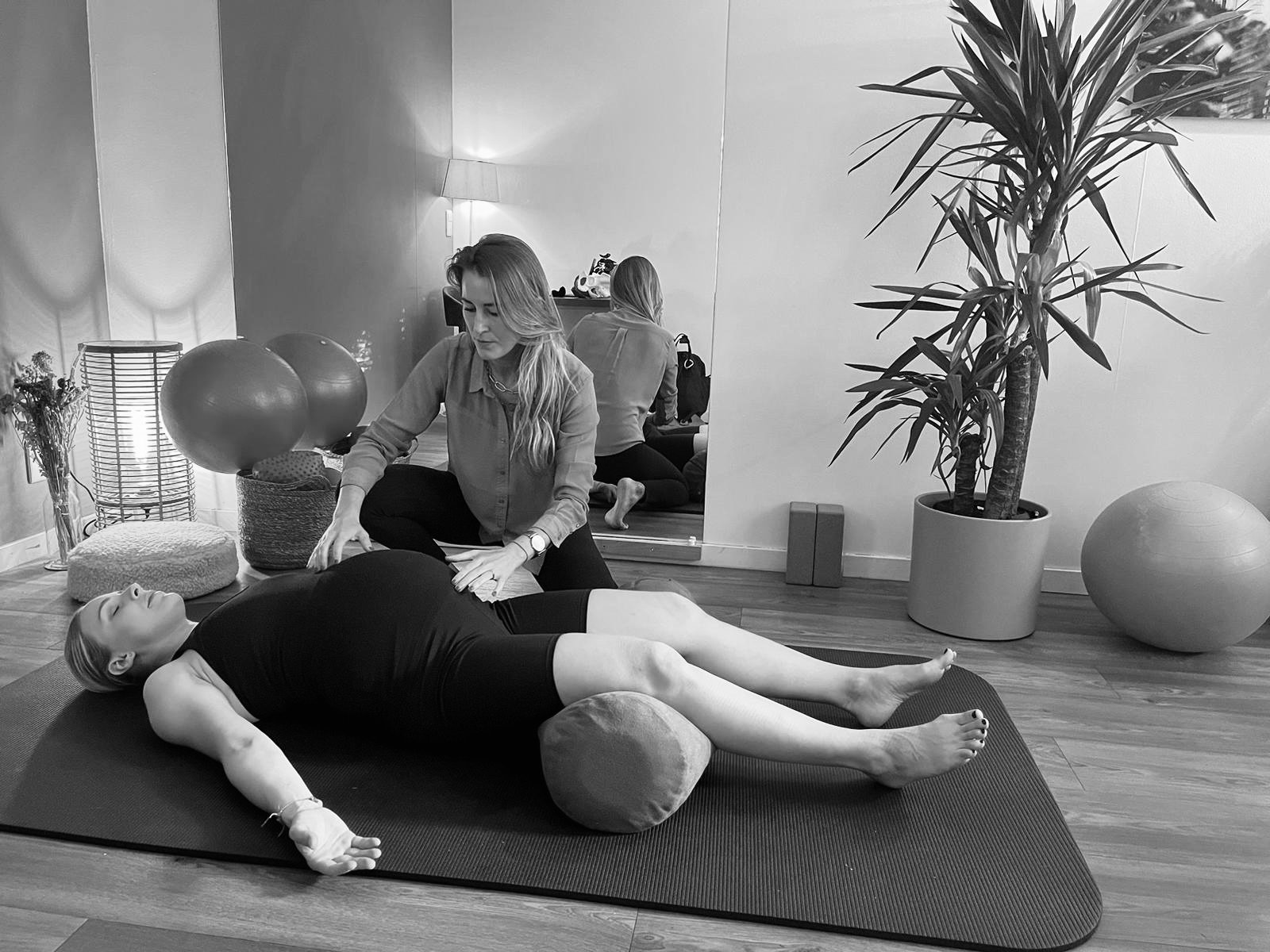 Deux femmes et un bébé lors d'une séance de kinésithérapie prénatale sur un tapis dans une salle lumineuse avec des plantes. femme guide l'autre qui s'étire avec le bébé posé devant elle. à Woluwe (Bruxelles)
