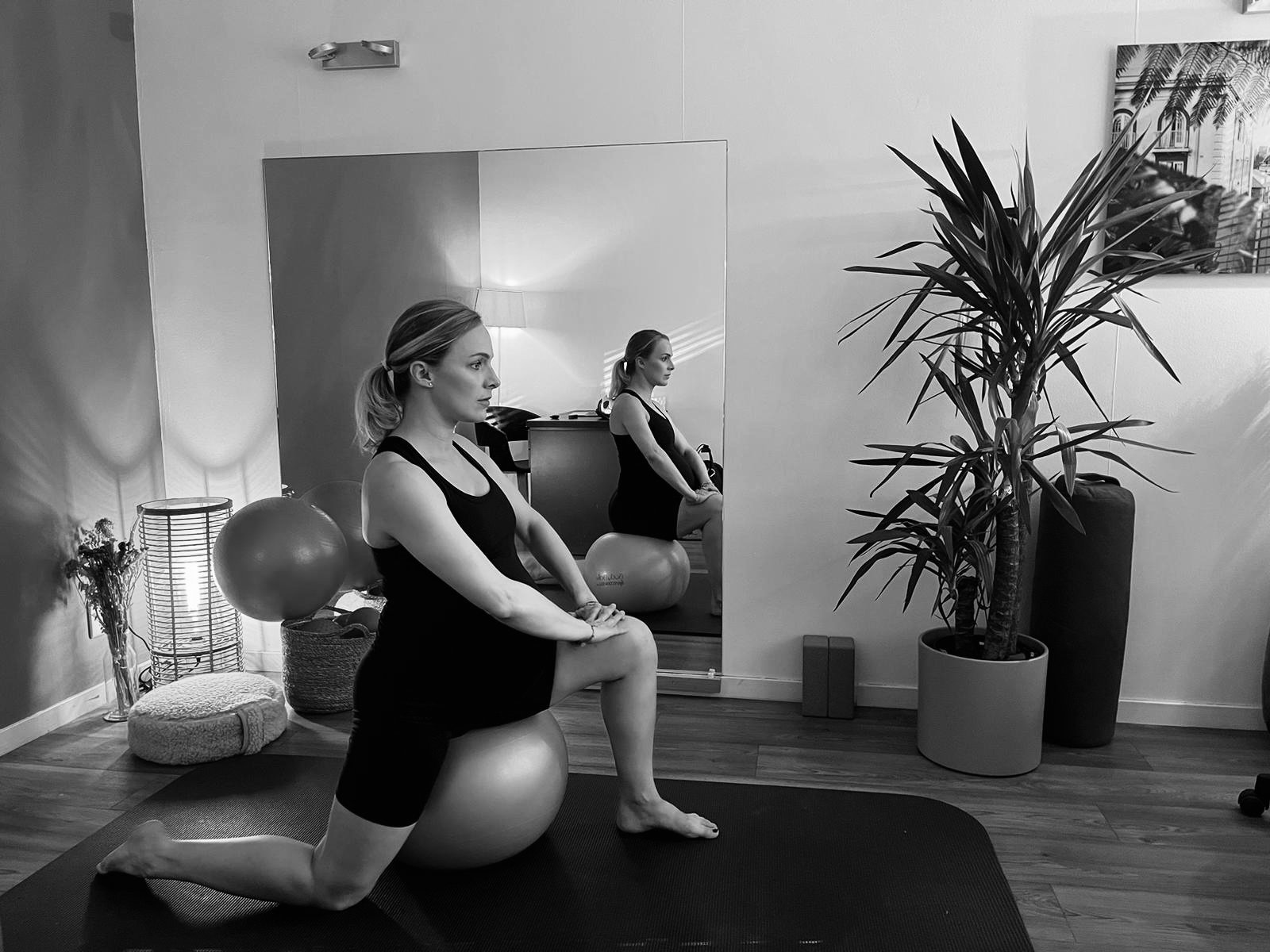 femme en tenue de sport s'étire sur un ballon de fitness dans une salle de sport, avec un miroir et des plantes en arrière-plan. à Woluwe (Bruxelles)