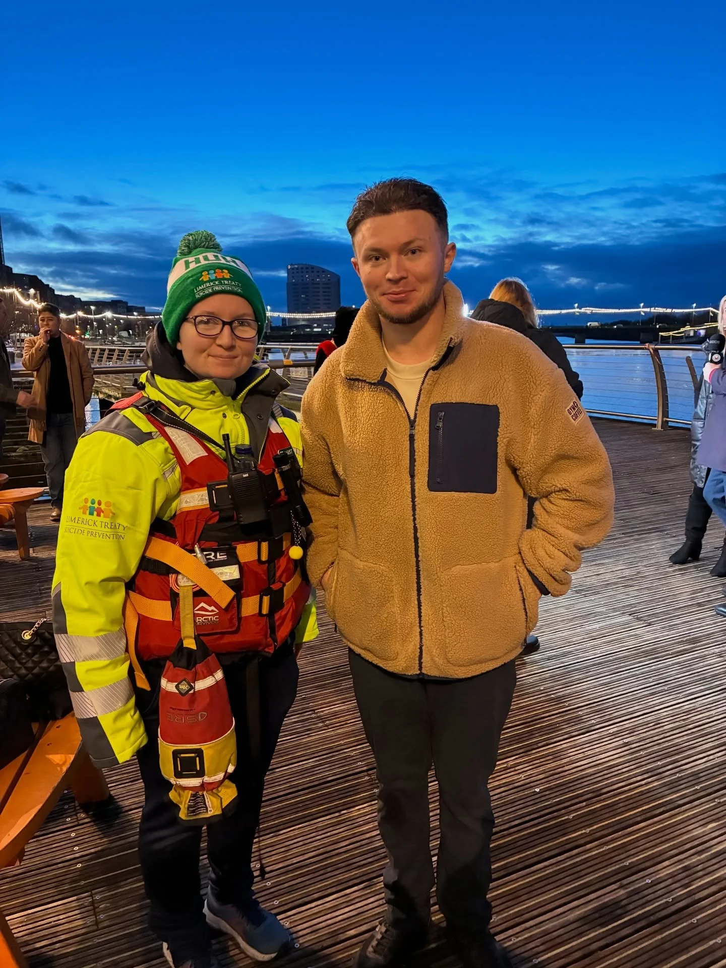 Two people standing on a wooden boardwalk near a river during dusk, with a city skyline and bridge in the background. One person wears a yellow emergency response jacket and hat, and the other wears a tan fleece jacket.