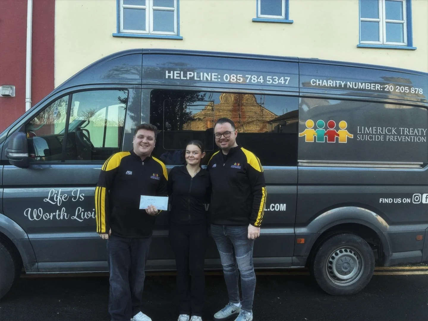 Three smiling people standing in front of a charity van, with two men and a woman. The van has charity logos, contact information, and a slogan that says "Life Is Worth Living." The van promotes Limerick Treaty Suicide Prevention.