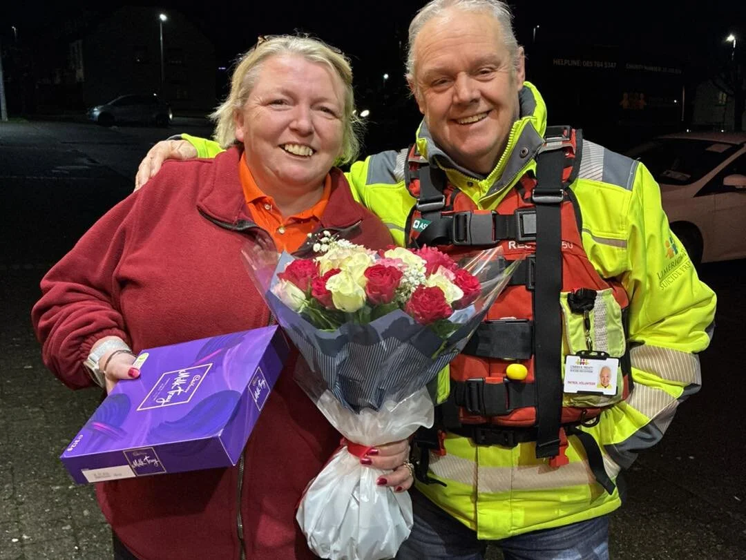 A woman holding a bouquet of pink and white roses and a box of chocolates standing next to a smiling emergency responder in a bright yellow and orange uniform at night.