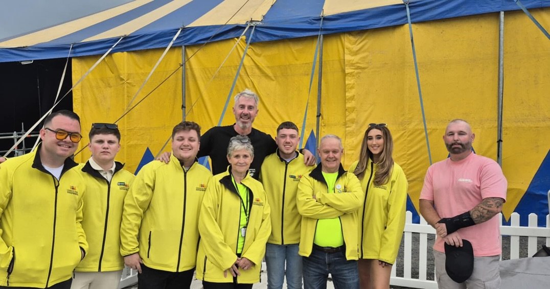 Group of eight people standing in front of a yellow and blue circus tent, wearing yellow jackets, with one man in a pink shirt on the right.
