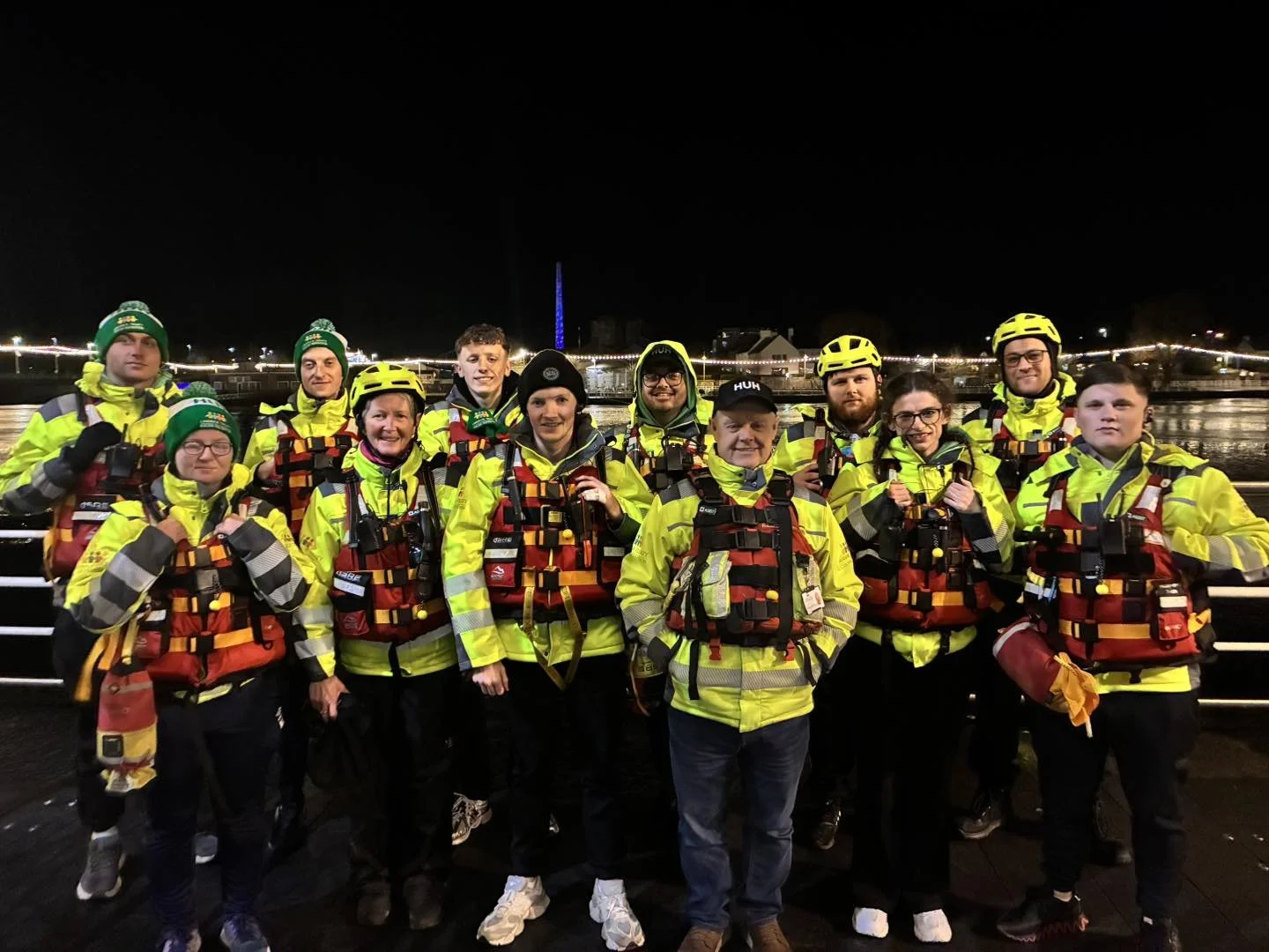 Group of people in yellow safety jackets and helmets standing on a boat at night with city lights and a lit-up Ferris wheel in the background.