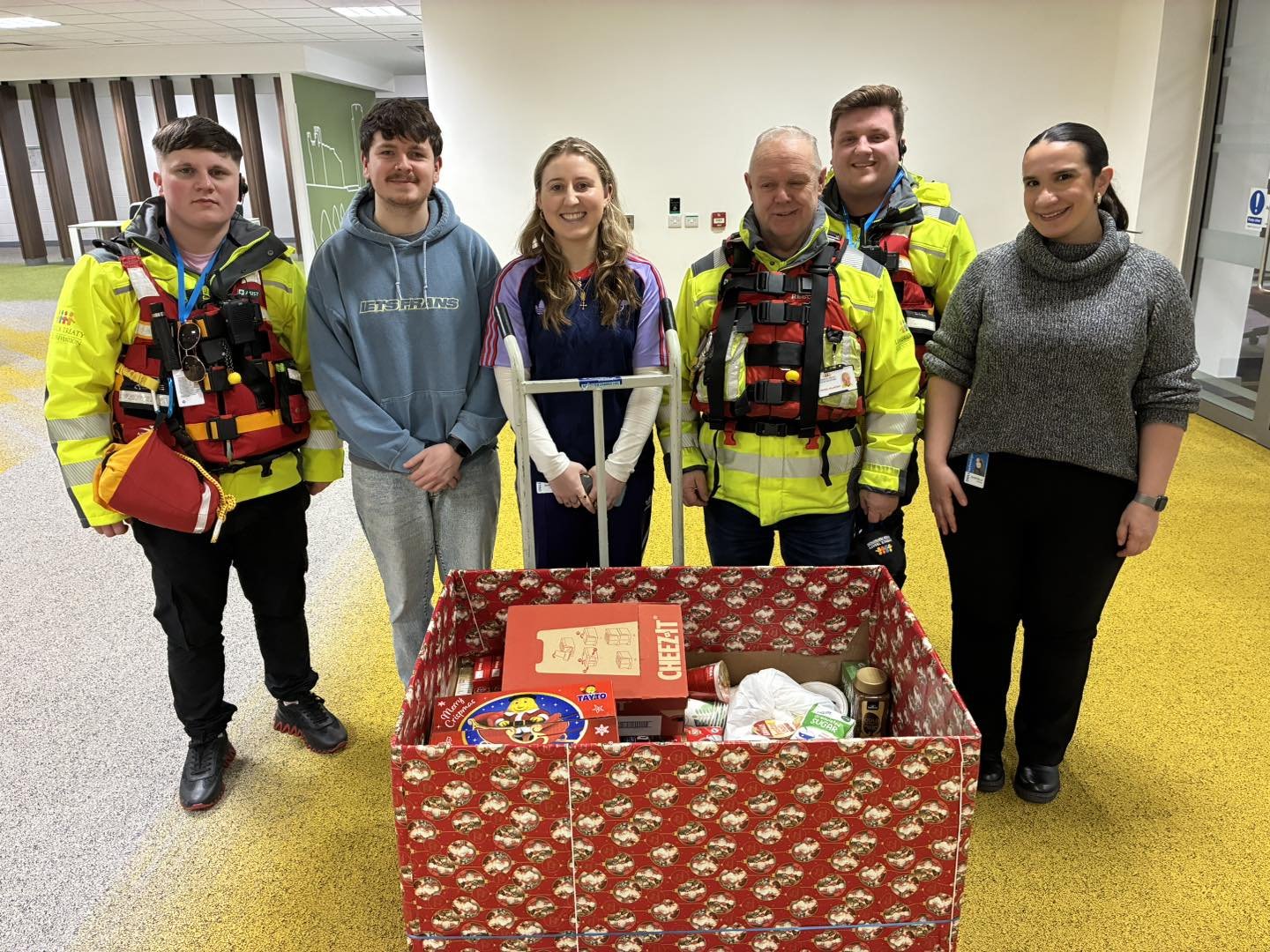 Group of six people standing behind a large red box filled with gifts and food, in an indoor setting, some wearing emergency responder uniforms.