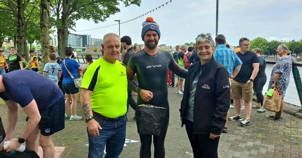 Three men, two of whom are wearing bright sportswear, stand together smiling near a river, with a crowd of people in the background, outdoors on a cloudy day.
