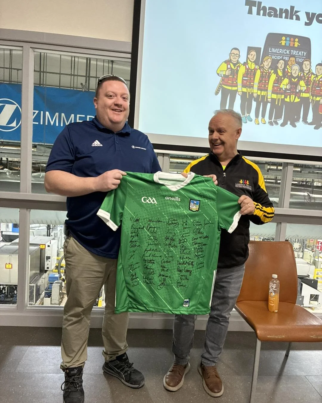 Two men standing indoors holding a signed green sports jersey, with a large screen displaying a cartoon firefighter group and the words "Thank you." There is a chair and a bottle of orange drink on the right side.