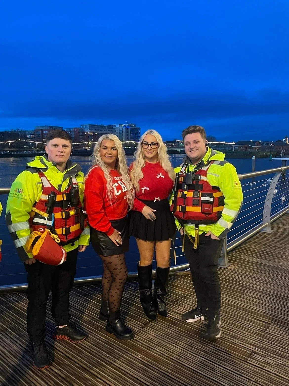 Four people standing on a wooden deck near a body of water during dusk, with city buildings in the background. Two are dressed in bright yellow and red emergency worker uniforms, and two women are dressed in holiday sweaters and outfits.