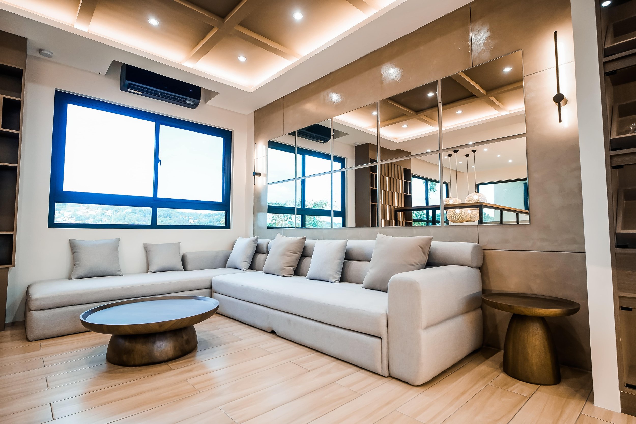 Modern living room interior with a beige sectional sofa, multiple beige cushions, a round wooden coffee table, mirrored wall panels, hardwood flooring, and a large window with natural light.