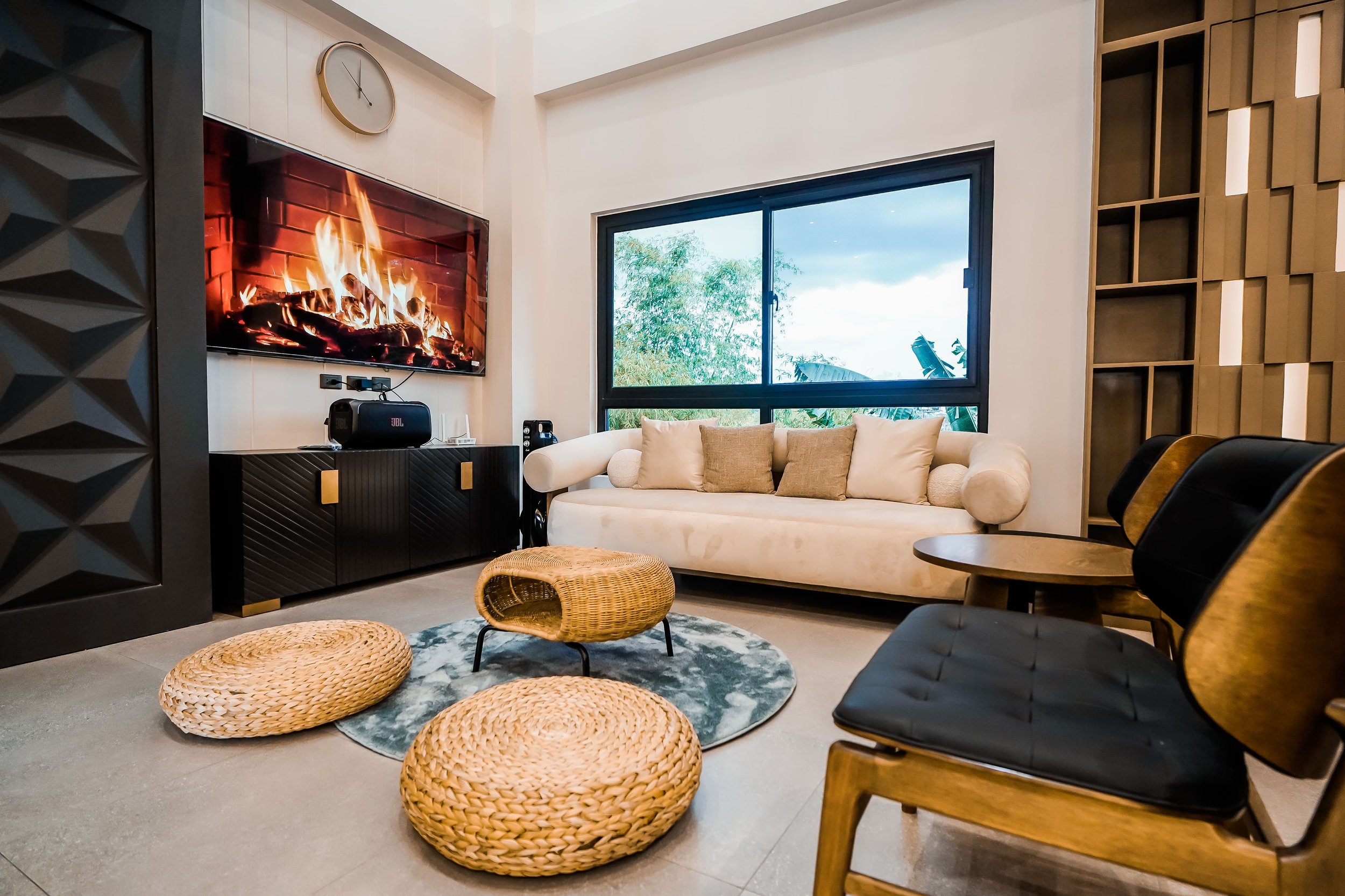 Modern living room with faux fireplace on TV, beige sofa, large window, woven ottomans, black cabinet, clock on wall, and contemporary decor.