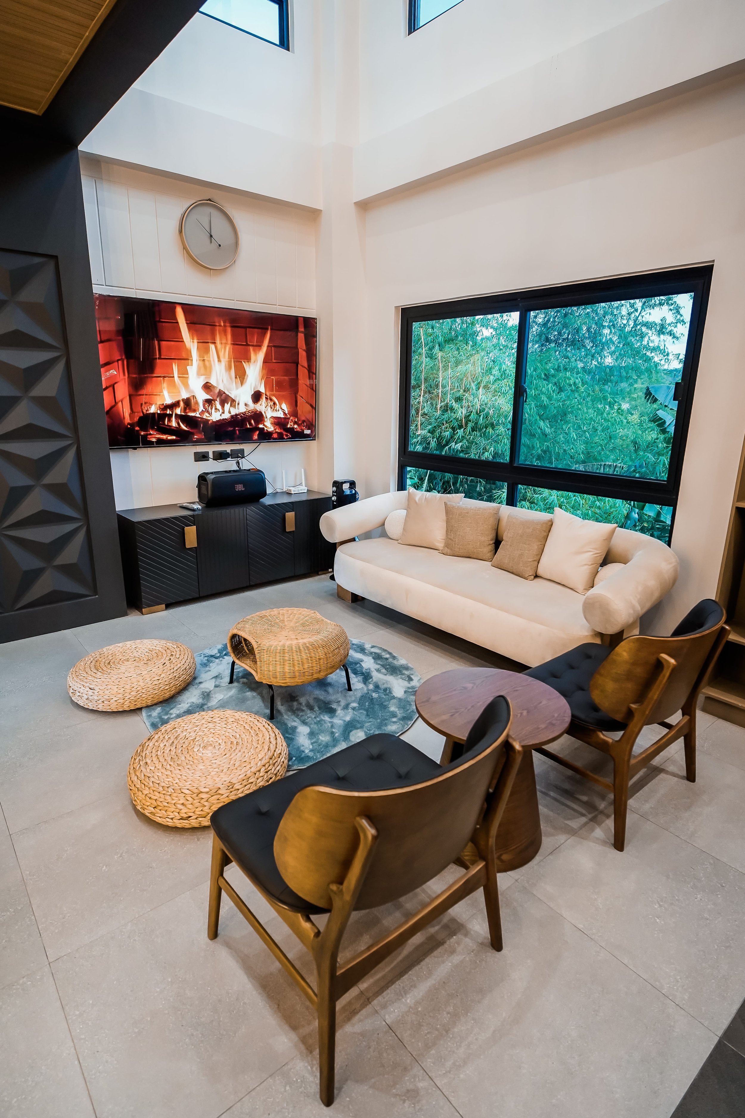 Modern living room with a mounted TV displaying a fireplace, a white sofa, two wooden chairs, a round side table, circular woven floor cushions, and a large window with a view of greenery.