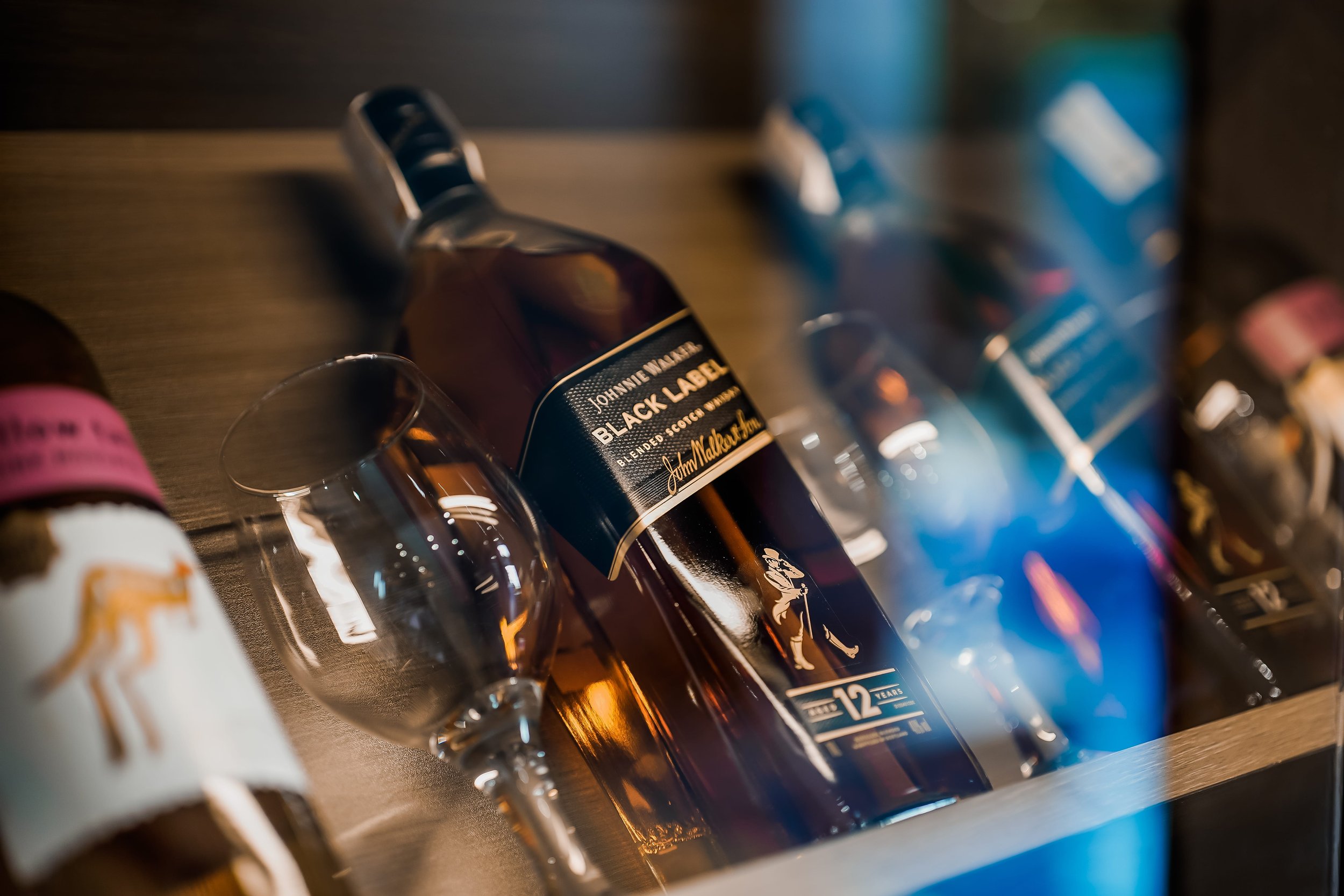 Close-up of a bottle of Johnnie Walker Black Label whiskey with a wine glass, surrounded by other bottles on a shelf.