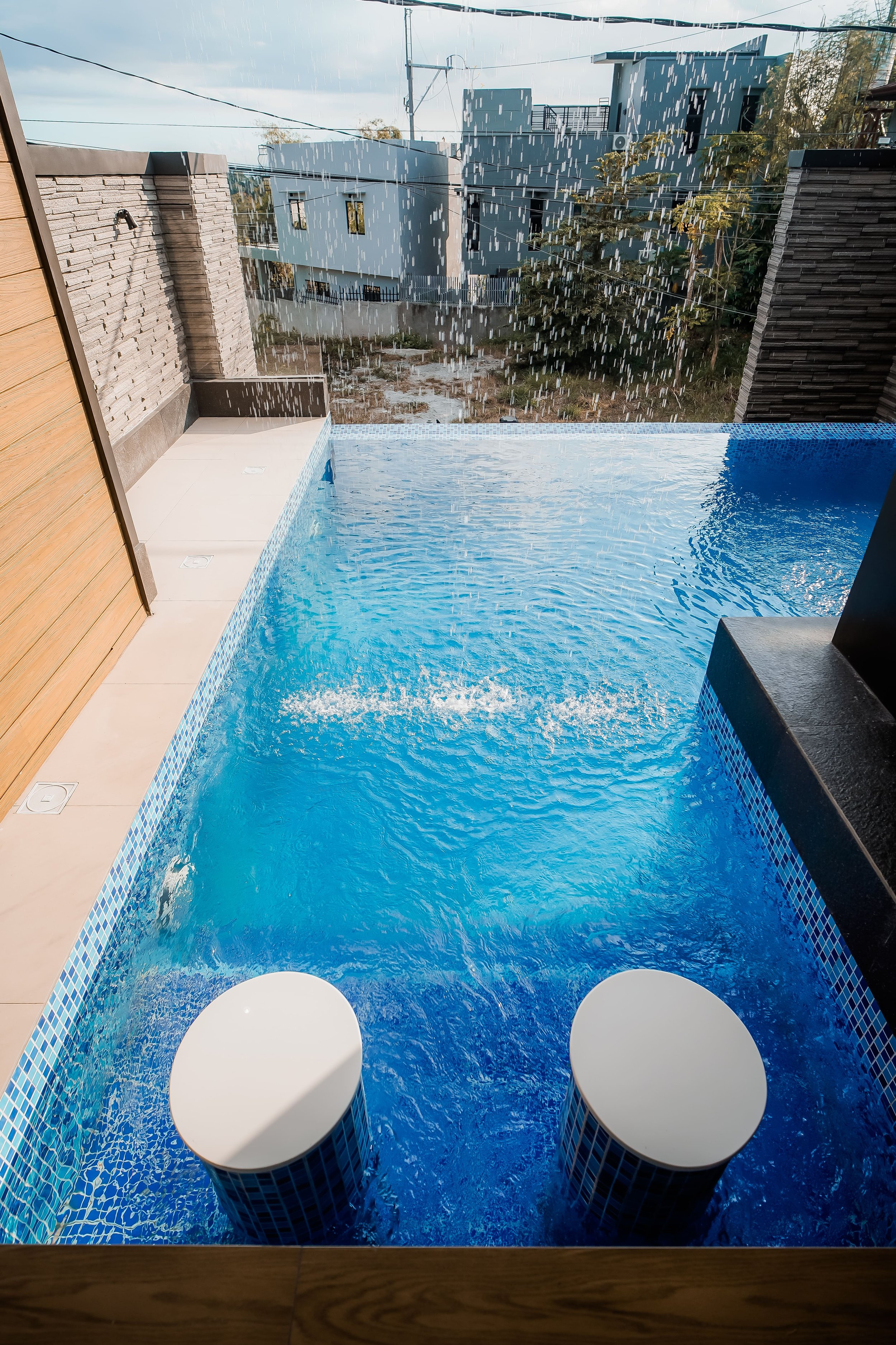 A modern outdoor swimming pool with water cascading down from above, featuring submerged round stools. The pool is bordered by a wooden deck and stone walls, with buildings visible in the background.
