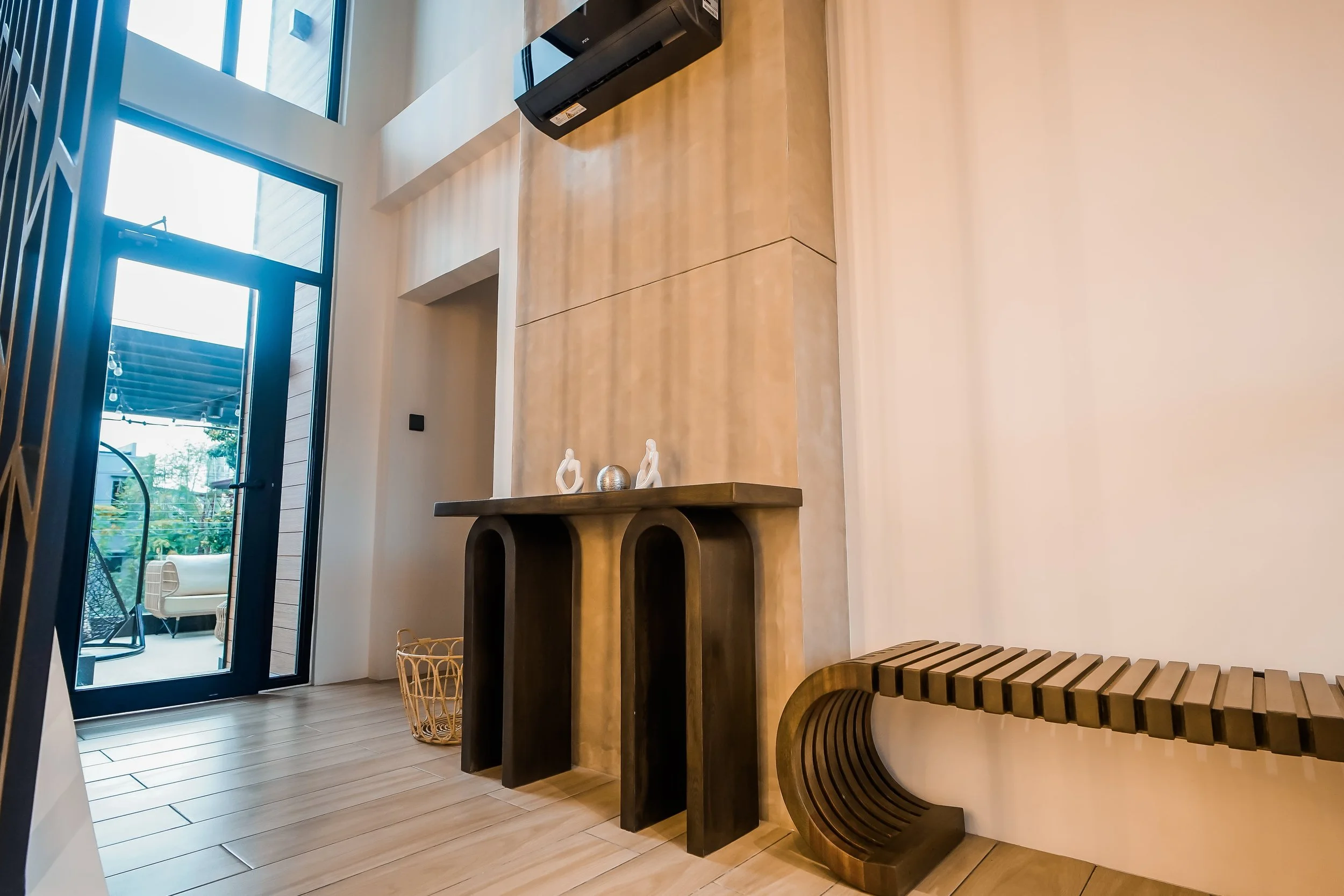 Modern living room interior with a wooden console table and decorative sculptures, a unique curved wooden bench, air conditioning unit, basket, and large windows leading to an outdoor area.