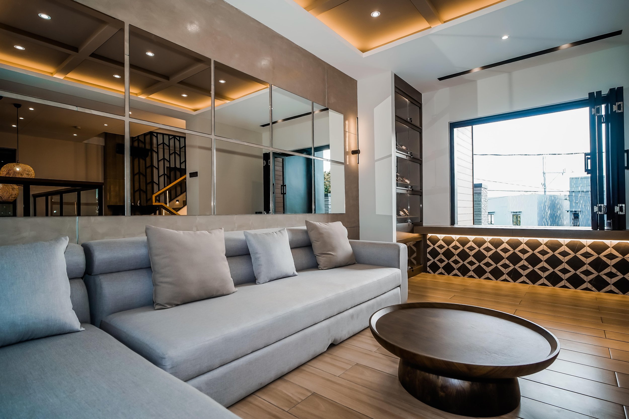 Modern living room with gray sectional sofa, decorative pillows, round wooden coffee table, geometric wall paneling, recessed lighting, large mirrors, and an open window view.