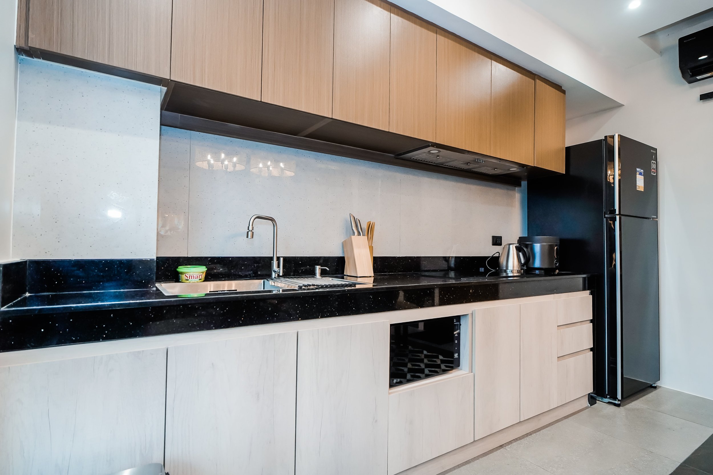 Modern kitchen with light wood cabinets, black countertop, stainless steel sink, knife block, electric kettle, and refrigerator.