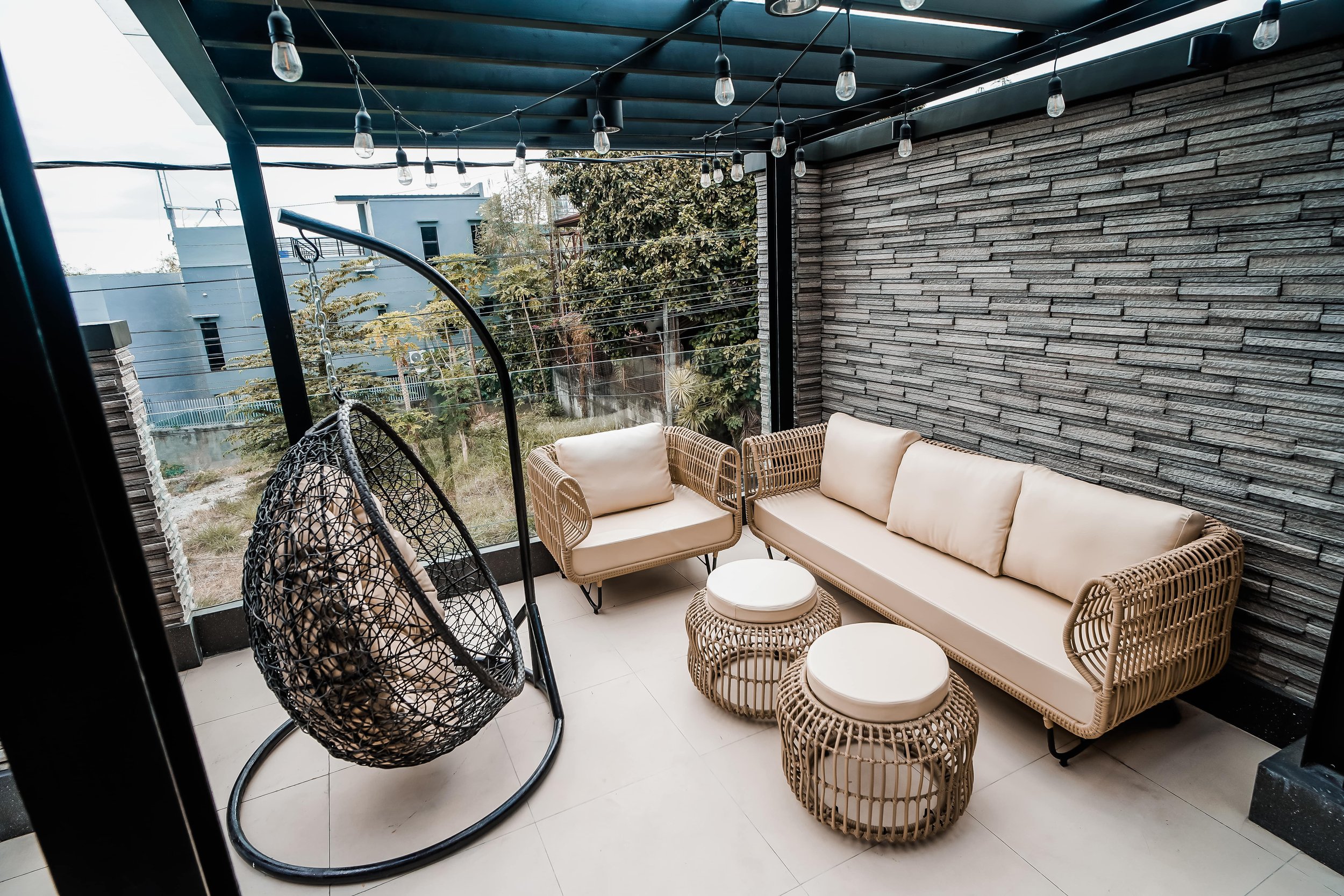 Outdoor patio with wicker furniture, hanging egg chair, brick wall, and string lights under a pergola.