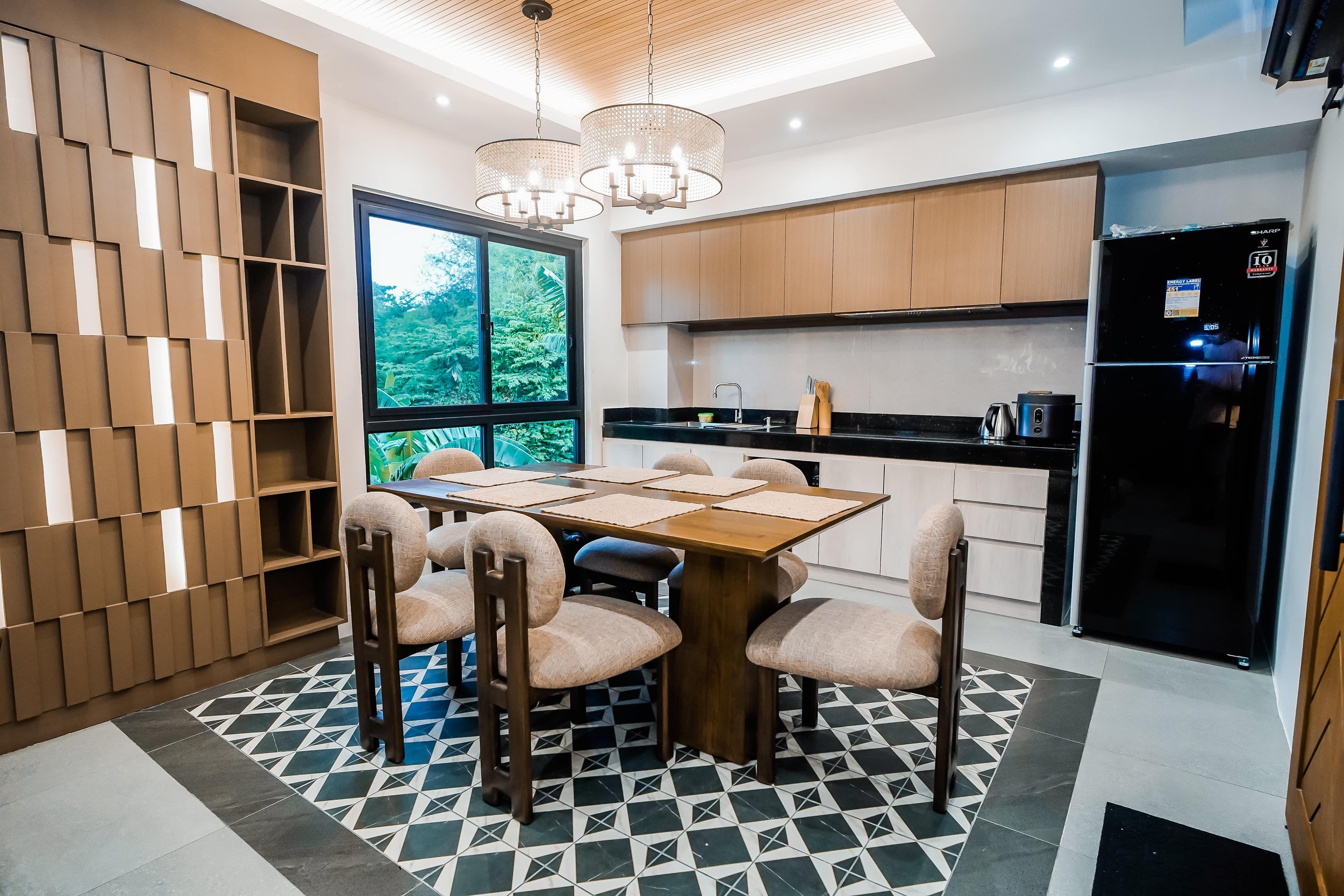 Modern kitchen and dining area with wooden cabinetry, a large window offering a green view, geometric tile flooring, a dining table with six chairs, and a black refrigerator.