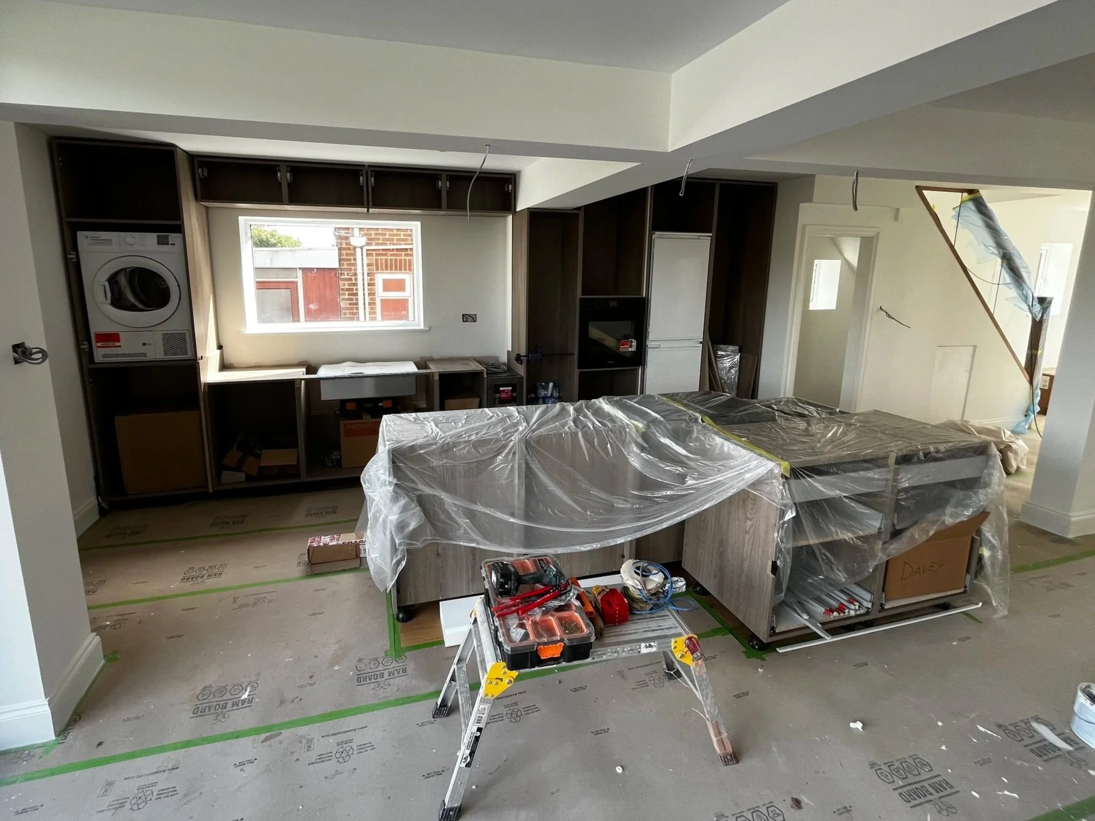 Kitchen under construction with wooden lower cabinets, white upper cabinets still in packaging, and a partially installed sink in a modern kitchen.