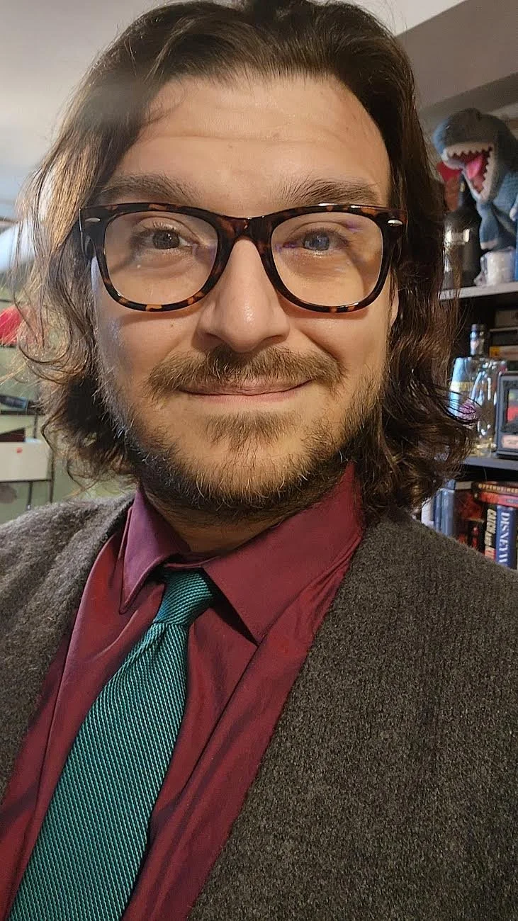 A man with long brown hair, glasses, and a beard, smiling at the camera. He's wearing a maroon shirt, a teal and black striped tie, and a brown blazer. Background includes shelves with books and a dinosaur figure.